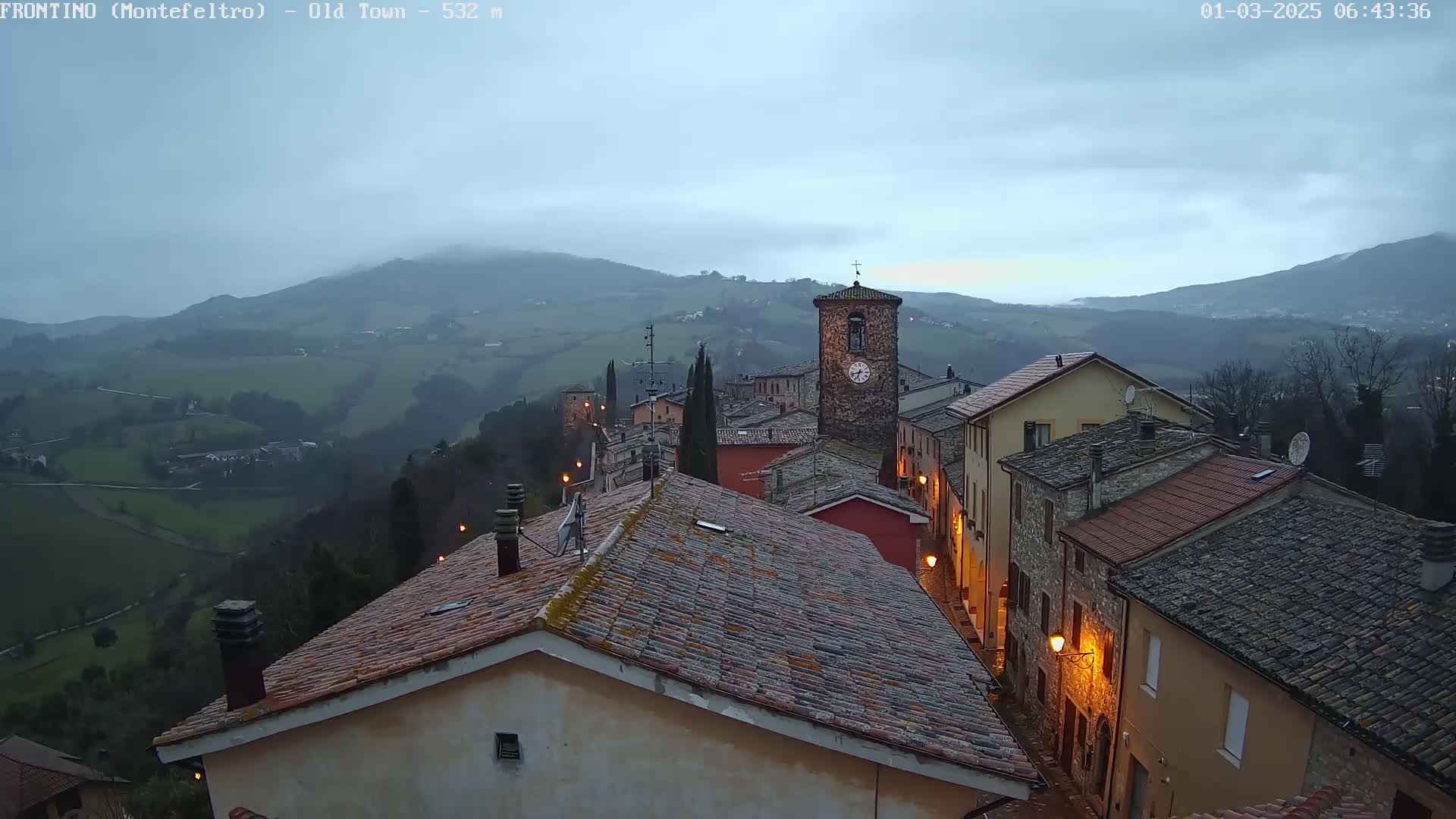 A stone village with a clock tower is nestled in a hilly landscape under an overcast sky.