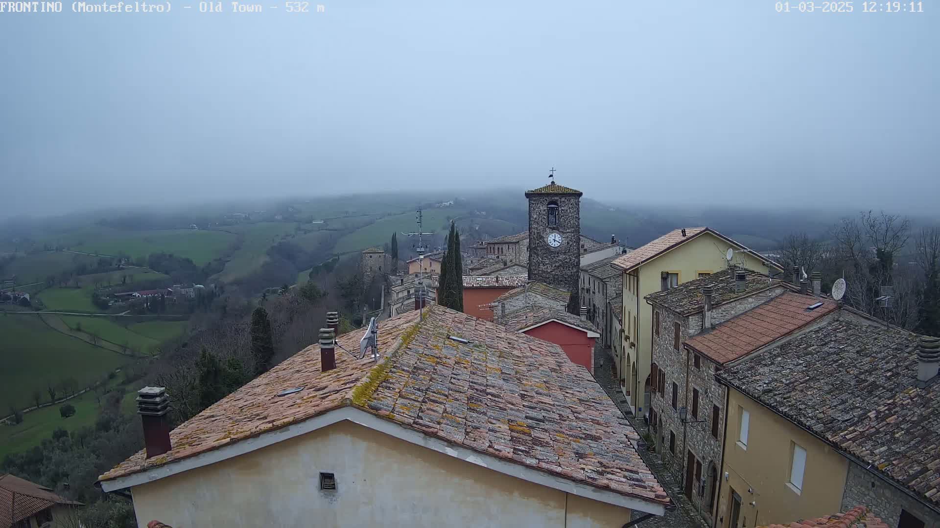 A stone village sits on a hillside overlooking a foggy valley of green fields.