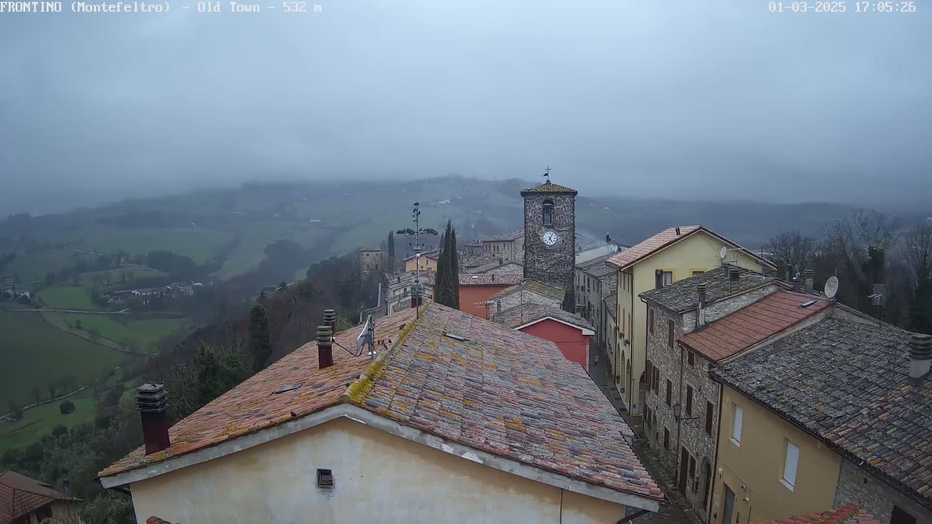 A stone village sits on a hillside under a foggy sky, overlooking a valley of green fields.