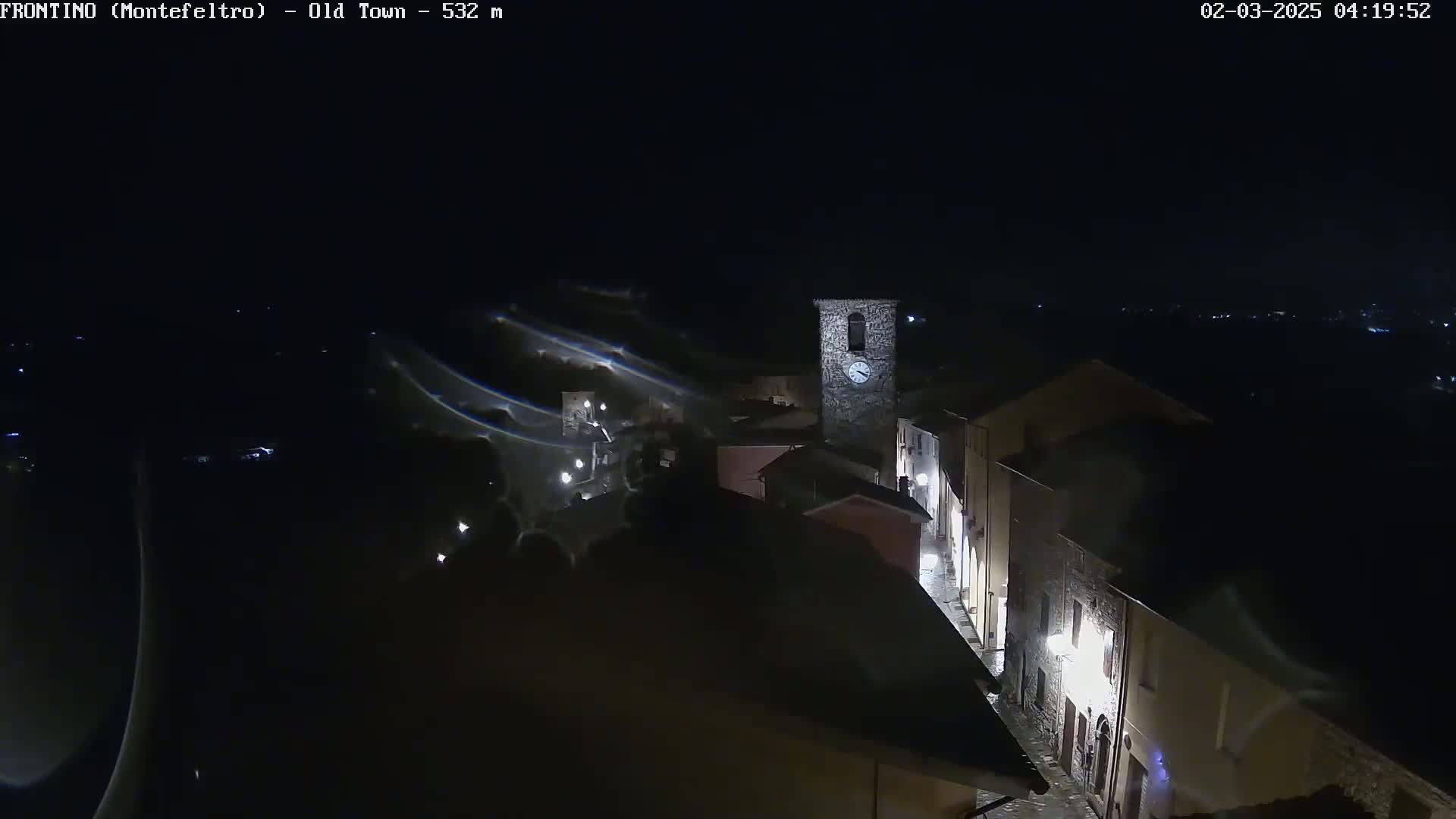 A nighttime, high-angle view of a small town's old section, featuring stone buildings illuminated by streetlights, with a clock tower prominently visible.