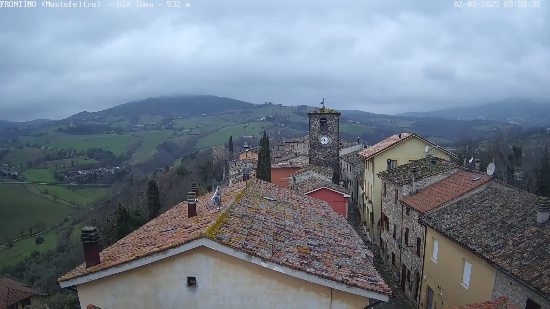 A view from a rooftop shows a small town nestled in a valley under an overcast sky, with rolling hills and mountains in the distance.