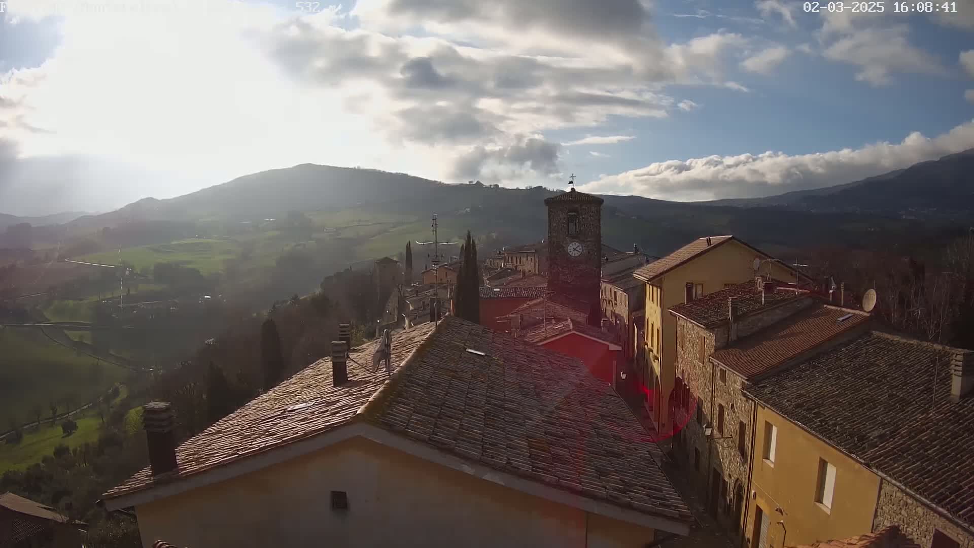 A partly sunny day reveals a quaint village nestled in a valley, surrounded by rolling hills under a partly cloudy sky.