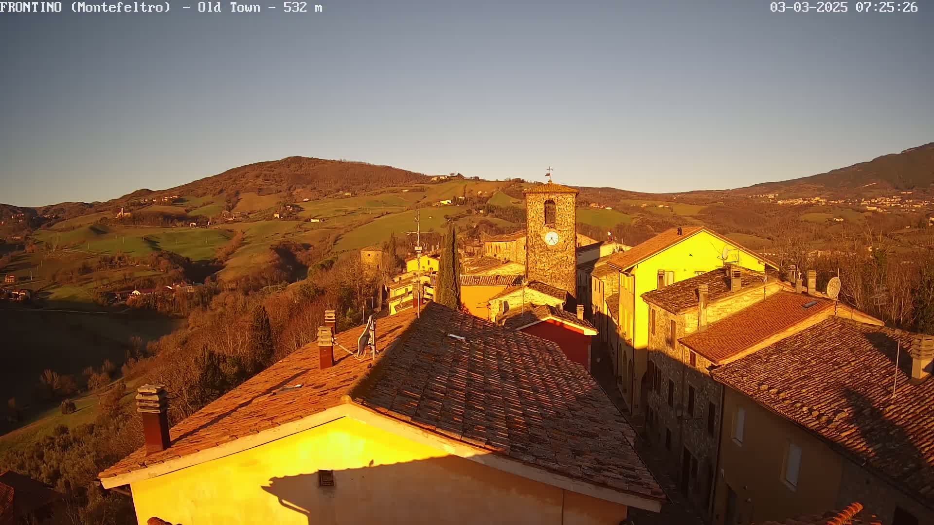 A sunlit hillside village with terracotta roofs and a clock tower is nestled in a valley, under a clear, pale blue sky.