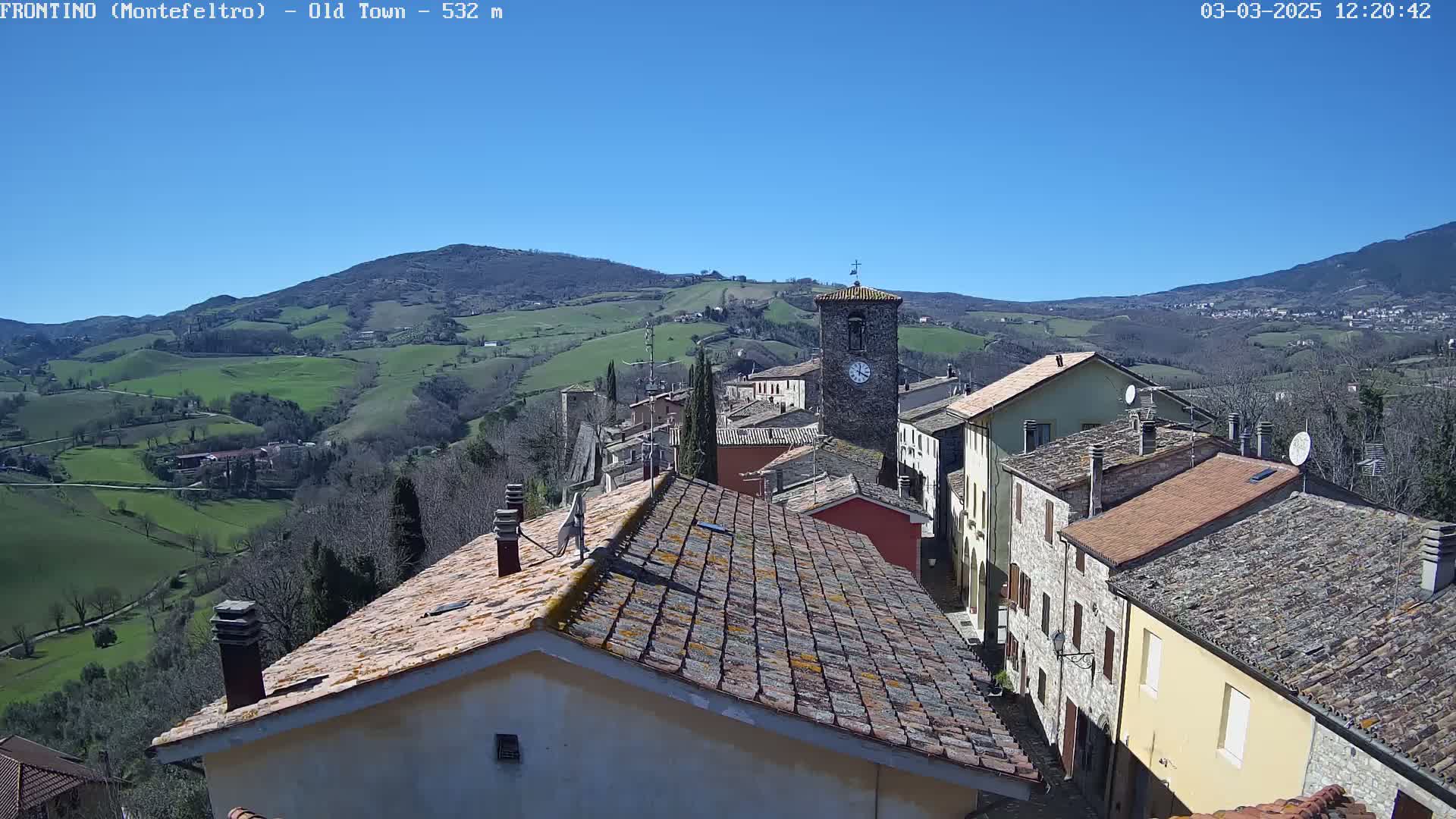 A stone village nestled in a valley under a clear blue sky, surrounded by rolling green hills.