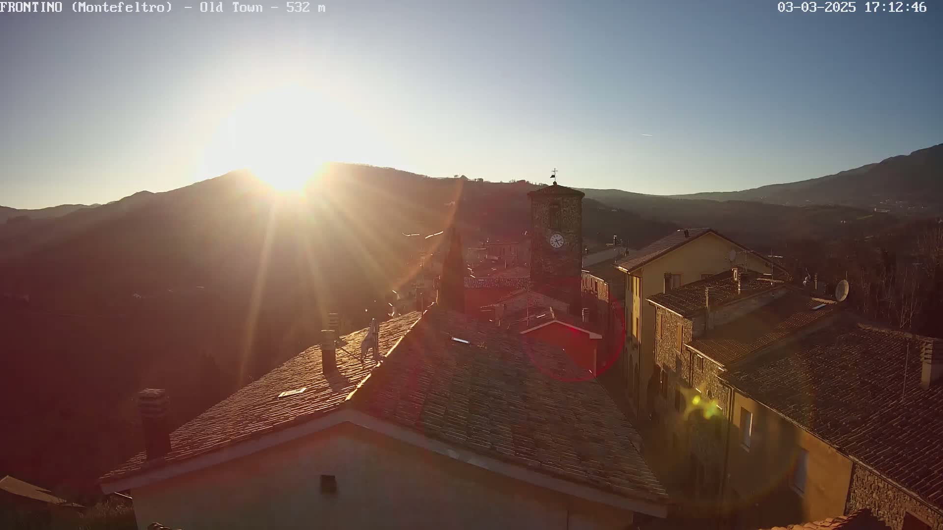 A sun-drenched, mountainous village with terracotta rooftops and a clock tower is seen at sunset.