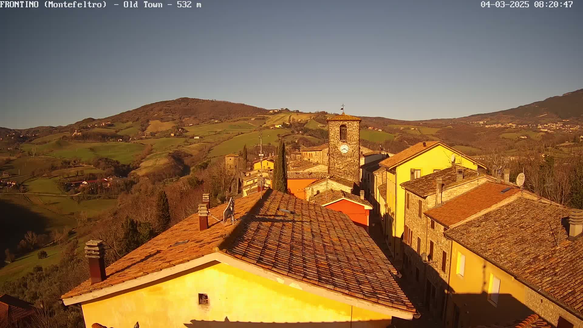 A sunlit village nestled in a valley between rolling hills, under a clear blue sky.