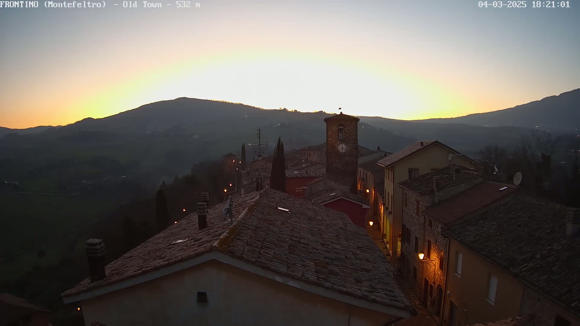 An elevated view of a small town nestled in a valley at sunset, with the last rays of light illuminating the rooftops and a clock tower.
