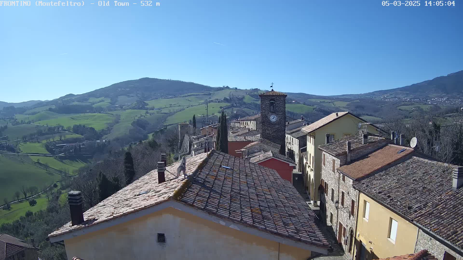 A clock tower and terracotta-roofed buildings sit atop a hill overlooking a sunny valley of rolling green fields.