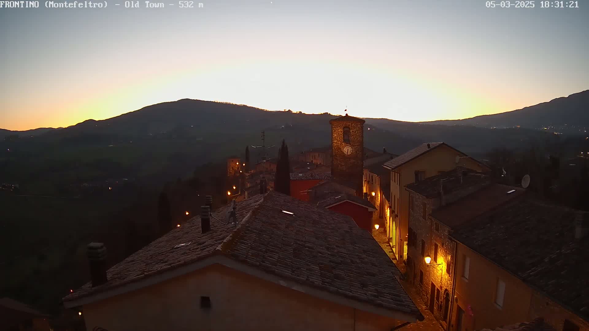 A hilltop village is illuminated at twilight, nestled among dark hills under a clear, dusky sky.