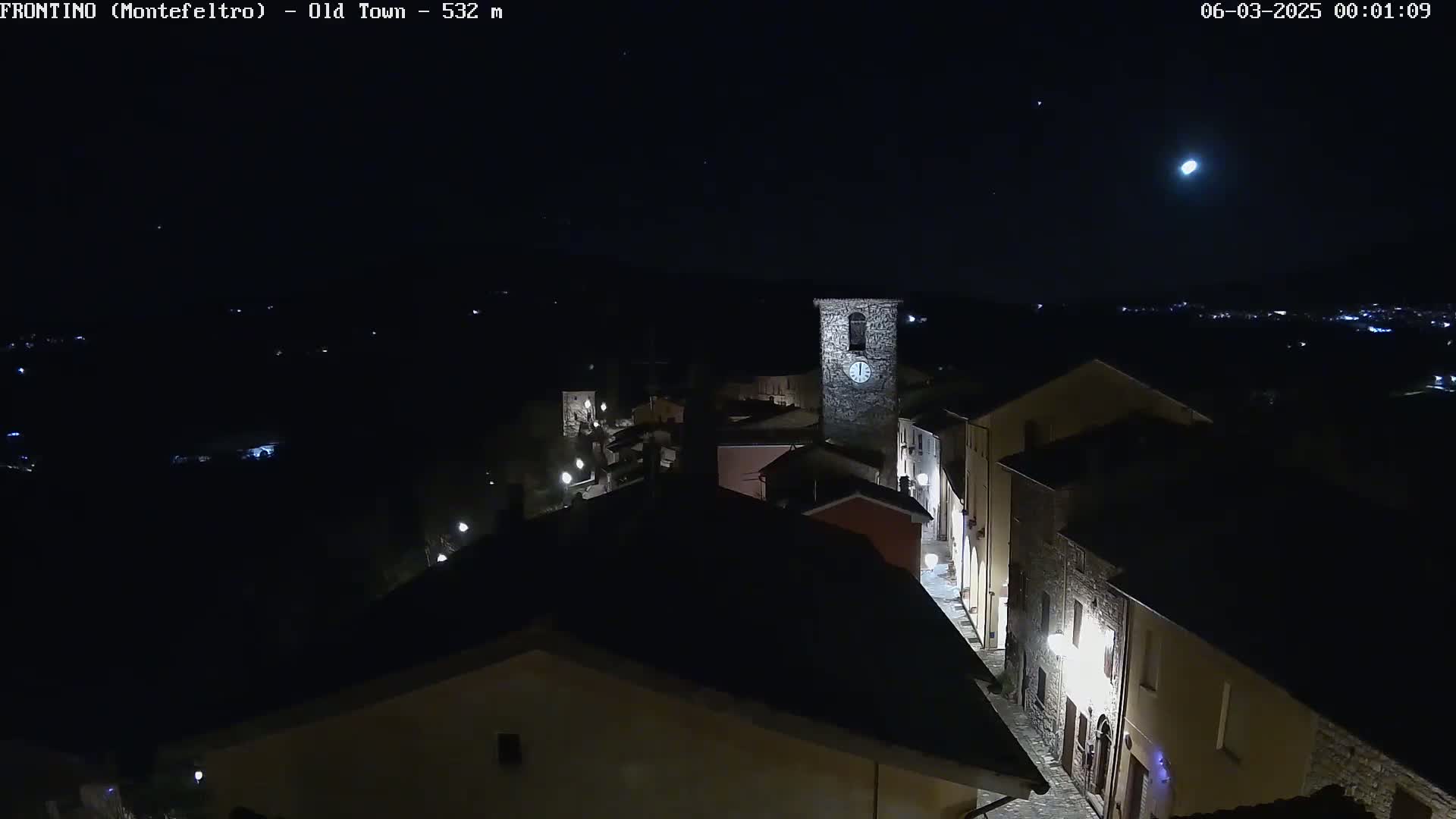 A nighttime, high-angle view of a small, stone village with a clock tower, illuminated by streetlights under a clear, moonlit sky.