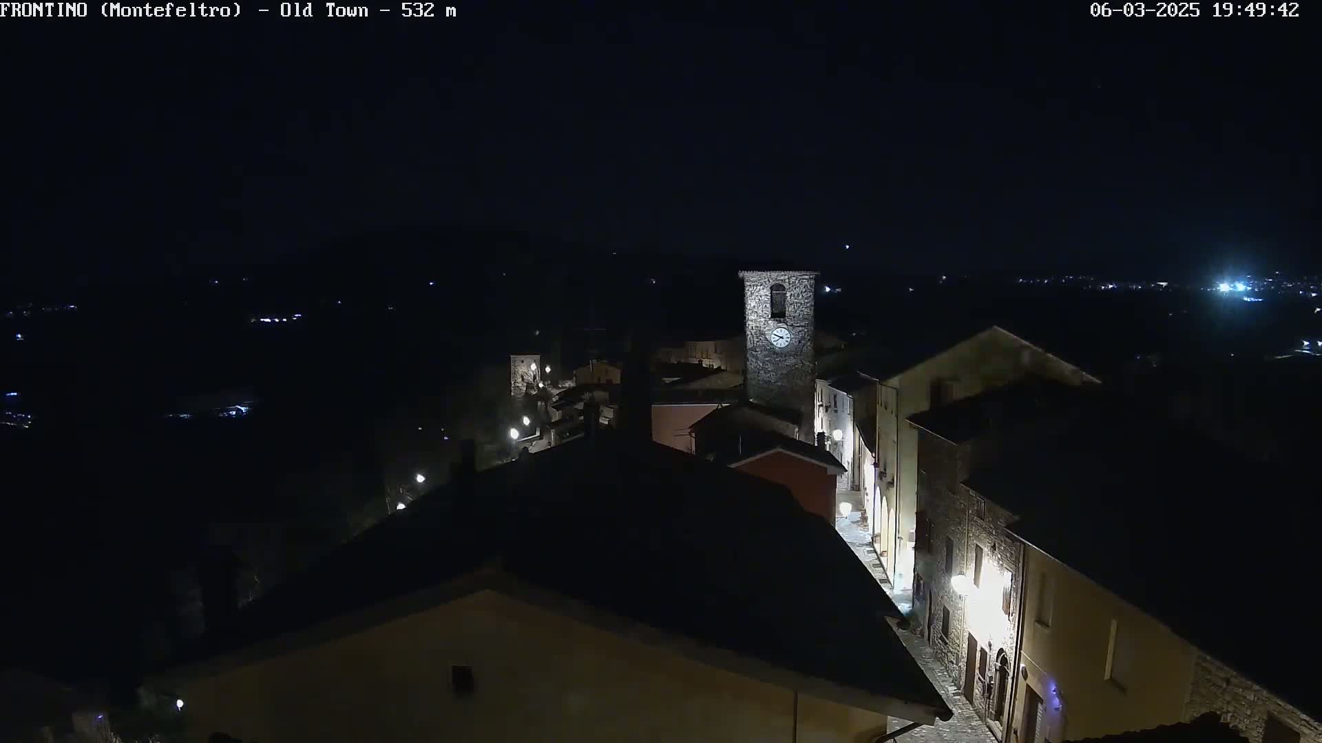 A nighttime, high-angle view of a dimly lit, stone village with a clock tower, situated on a hill overlooking a dark valley.