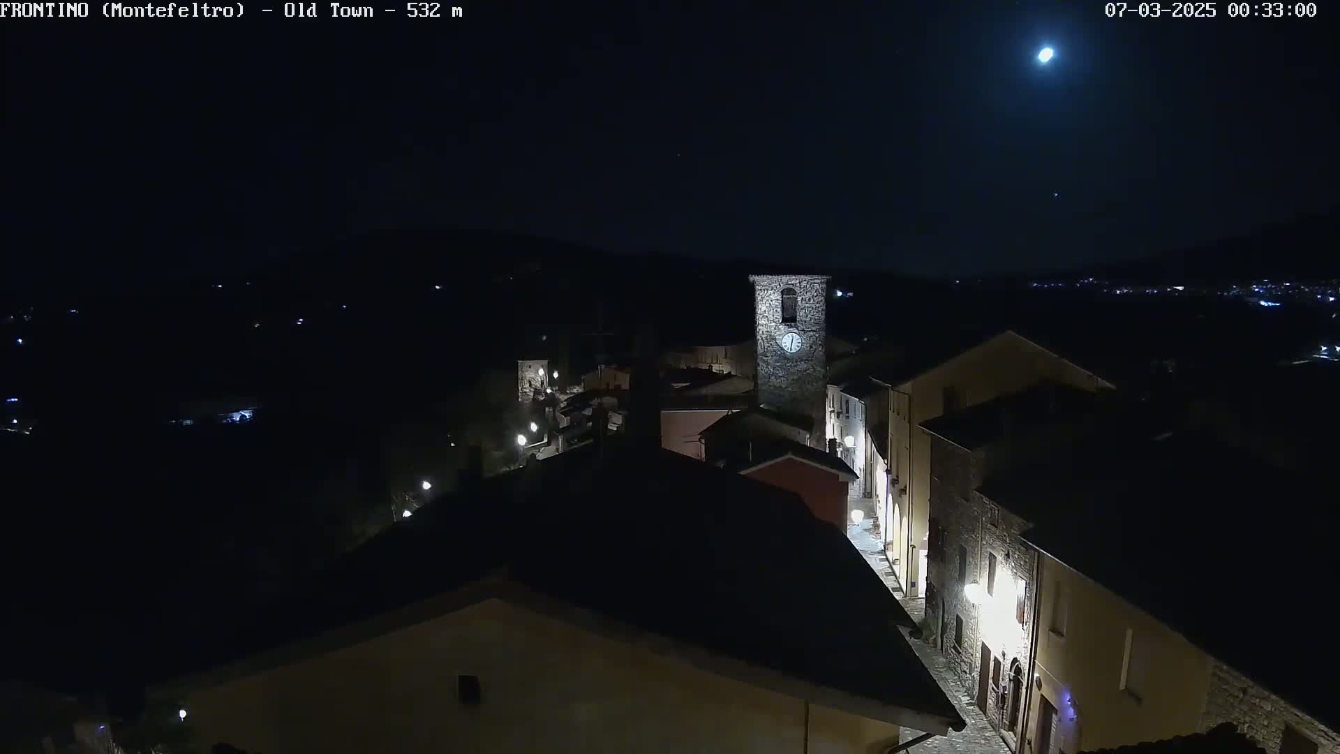 A nighttime, high-angle view shows a small town with a clock tower, illuminated by streetlights under a bright moon and clear sky.