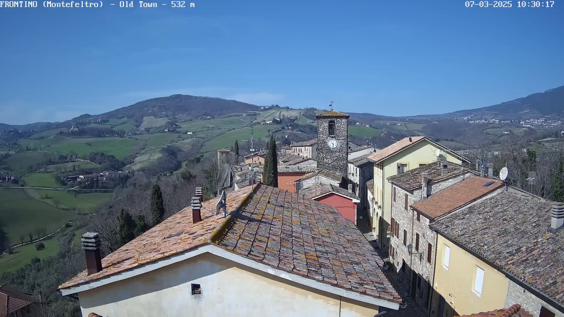 A small town nestled in a valley on a sunny day, with rolling green hills and mountains in the background.