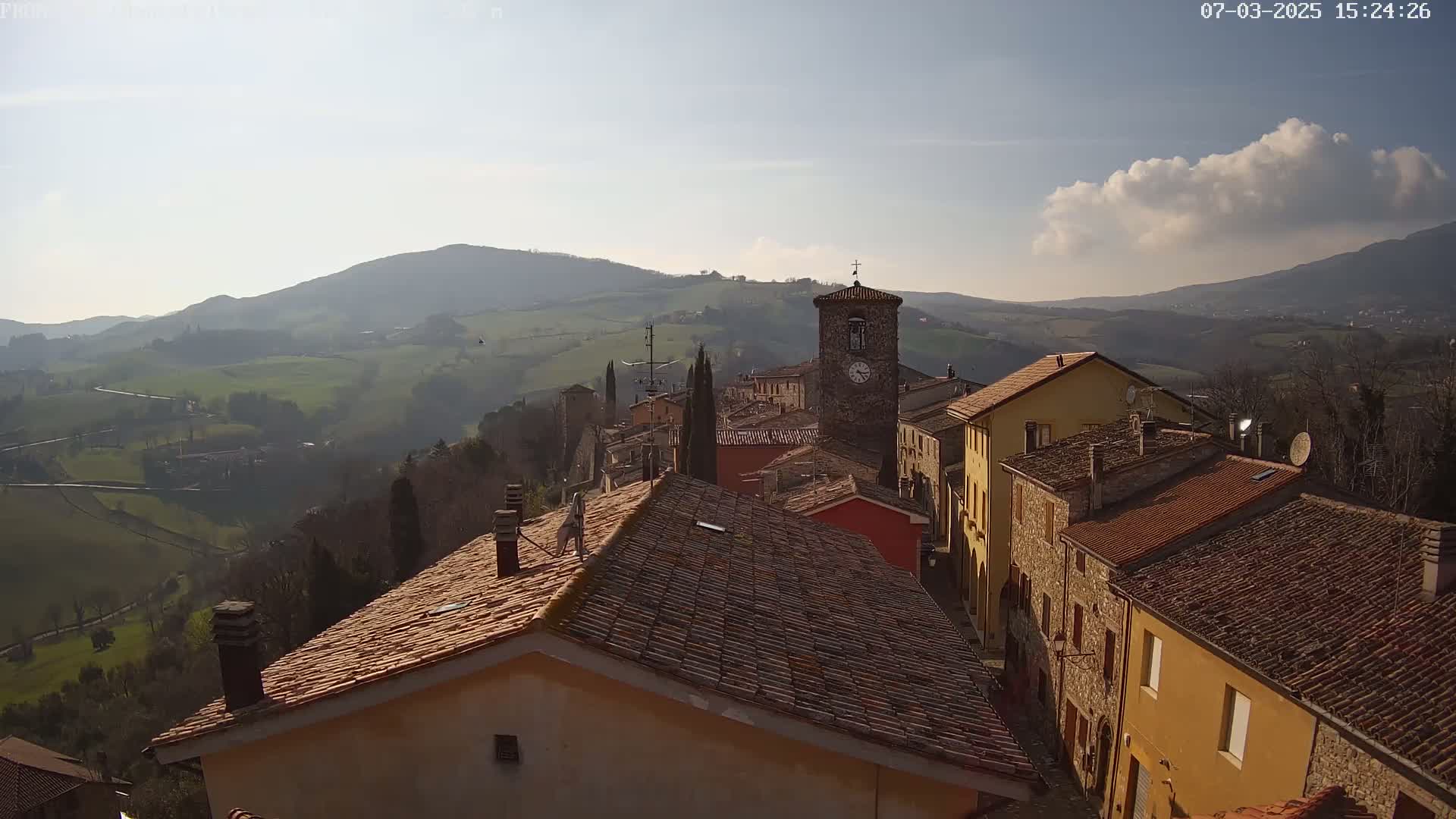 A sunlit, hilly landscape overlooks a small village of terracotta-roofed buildings, one of which has a clock tower, under mostly clear skies with a single, puffy cloud.