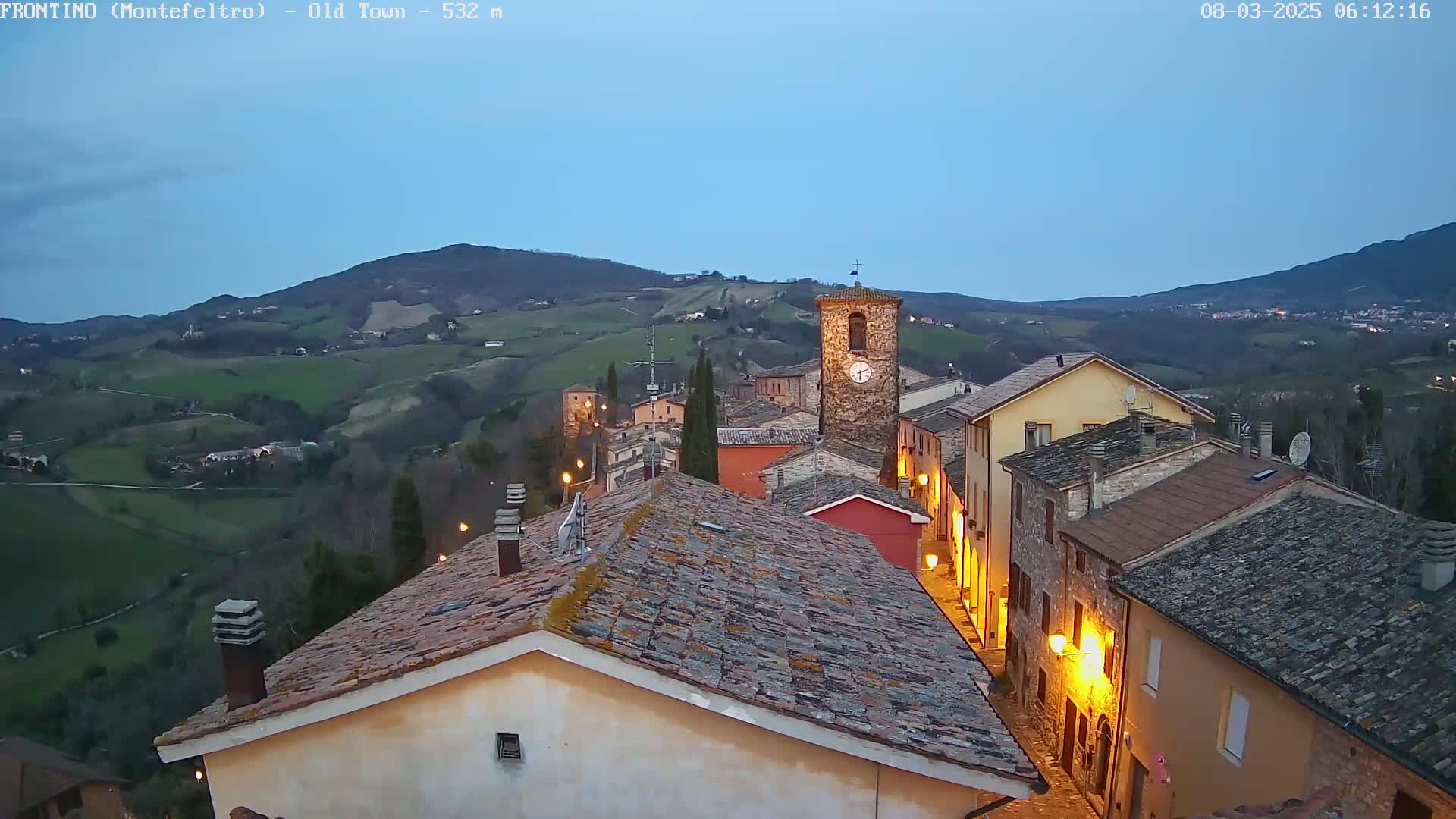 An Italian village nestled in rolling hills under a twilight sky is illuminated by streetlights.
