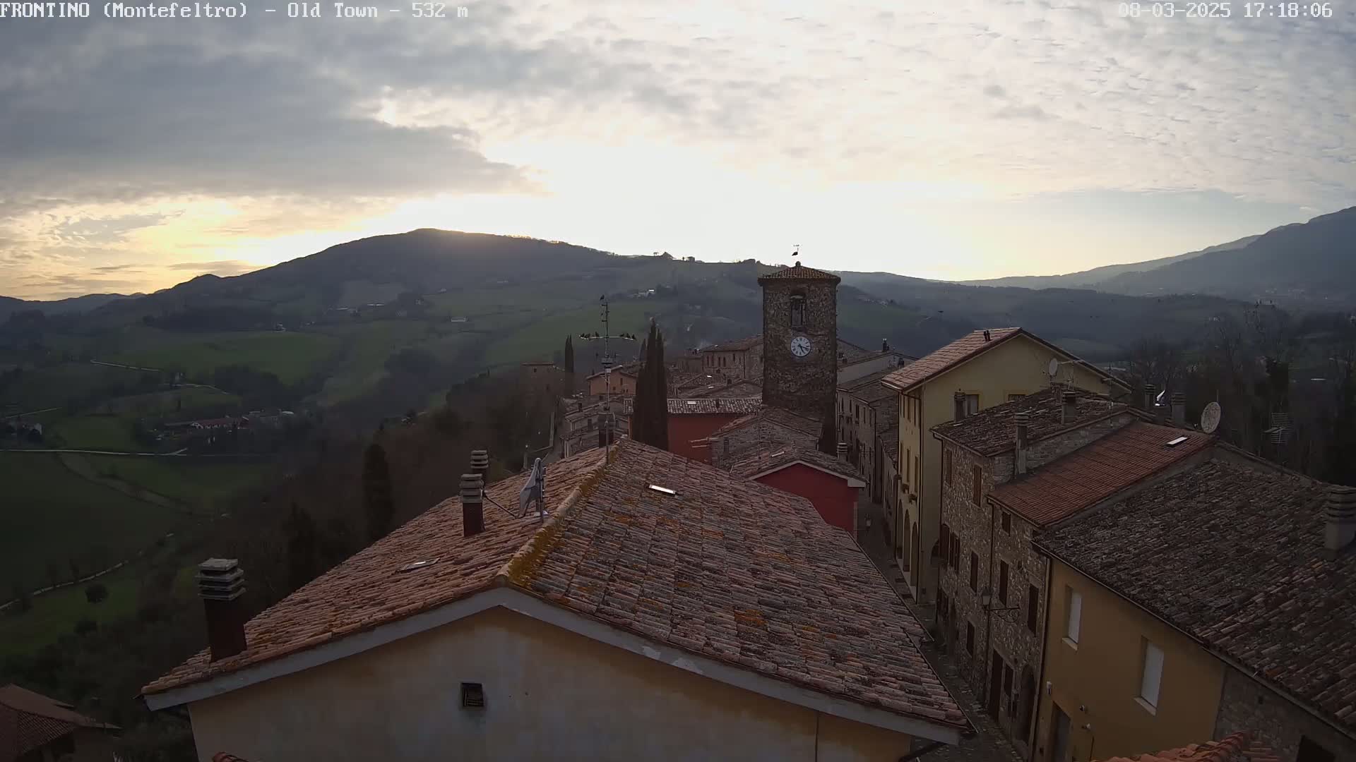 A stone village nestled in a valley, under a partly cloudy sunset sky, overlooks rolling hills and distant mountains.