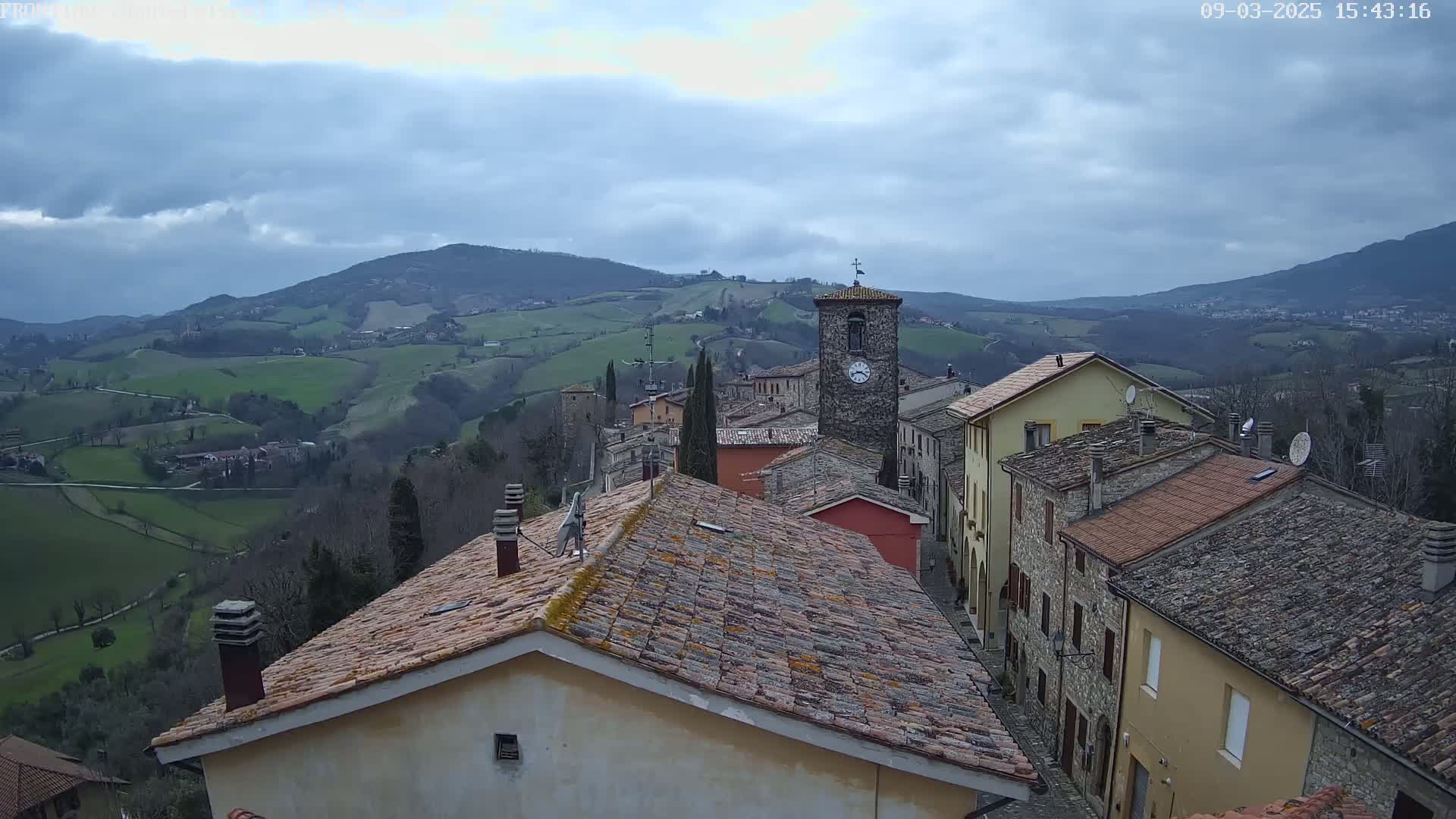 A stone village with a clock tower is nestled in a hilly landscape under a cloudy sky.