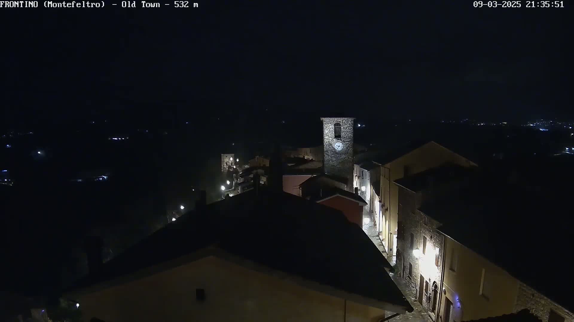 A nighttime, high-angle view shows a small, dimly lit town with a clock tower, under a dark sky.