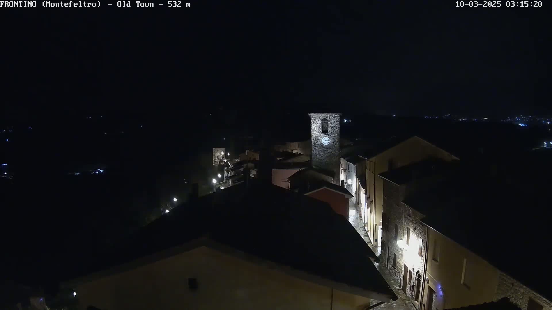 An aerial nighttime view shows a small town with dimly lit buildings and streets, a clock tower, and a dark sky.