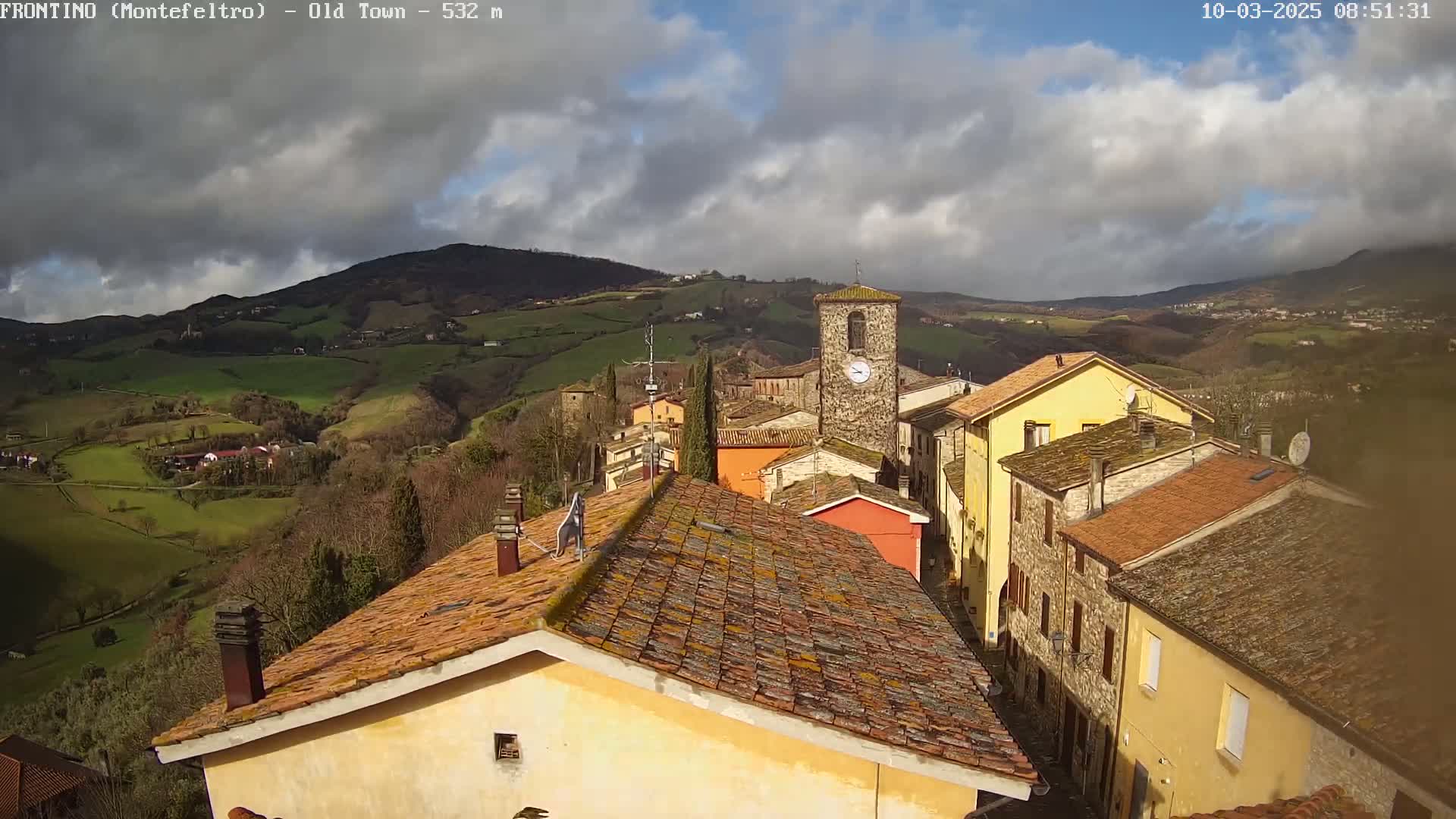 A partly cloudy day reveals a village nestled in a valley, with rolling green hills and mountains in the background.
