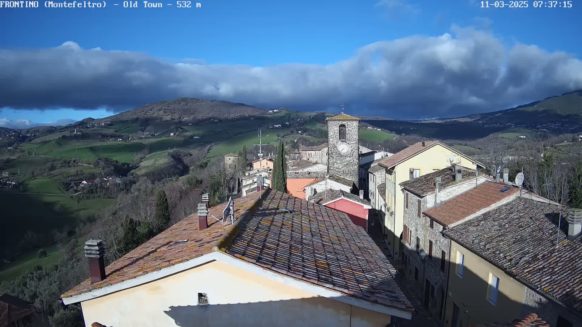 A view of a small town nestled in rolling hills under a partly cloudy blue sky.