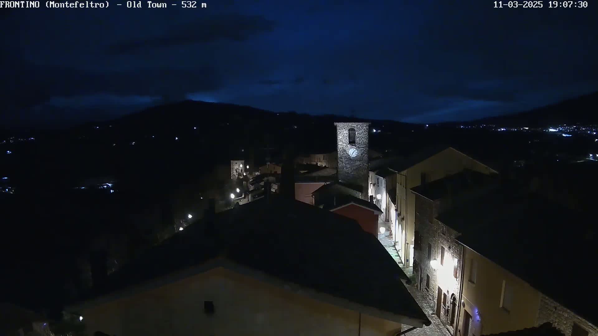 A nighttime, high-angle view shows a small, dimly lit town nestled in a valley under a dark, cloudy sky.