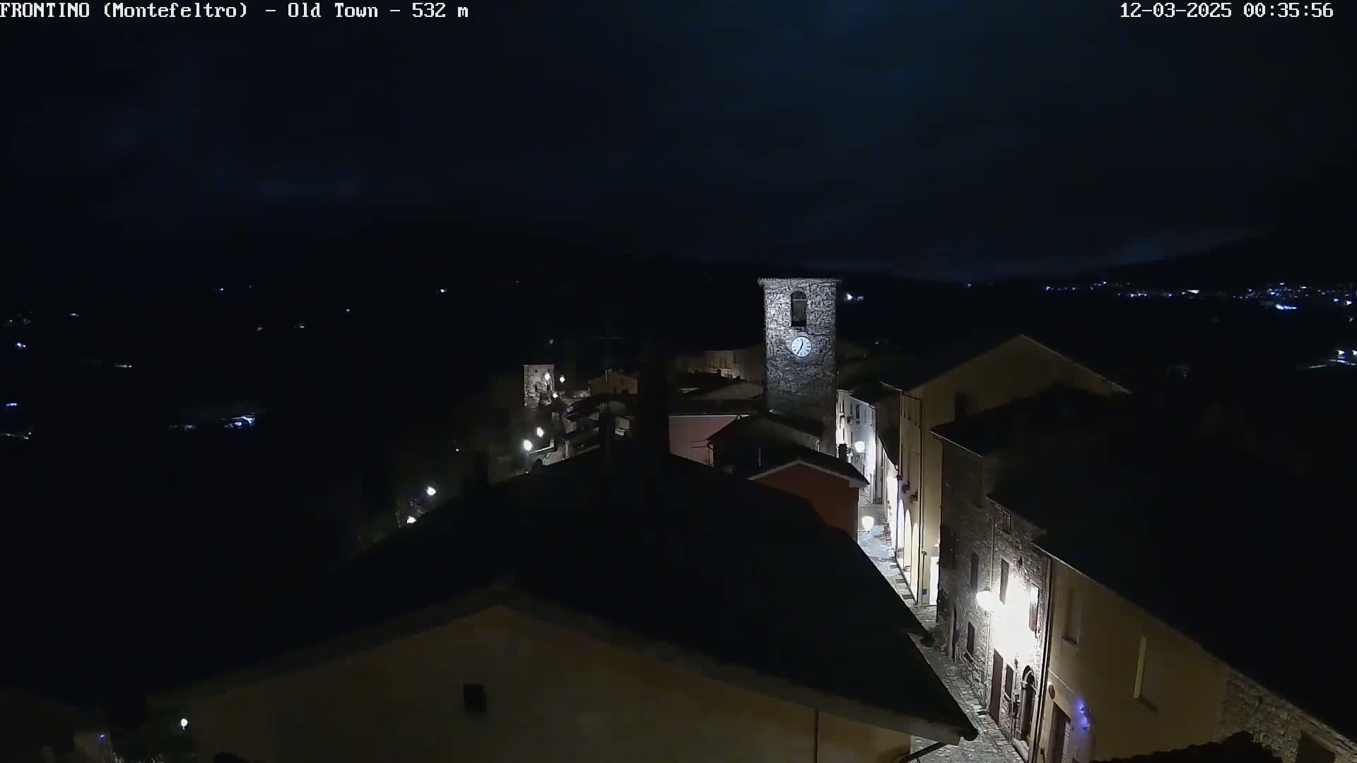 A nighttime, high-angle view of a small, stone town with a clock tower, illuminated by streetlights under a dark sky.
