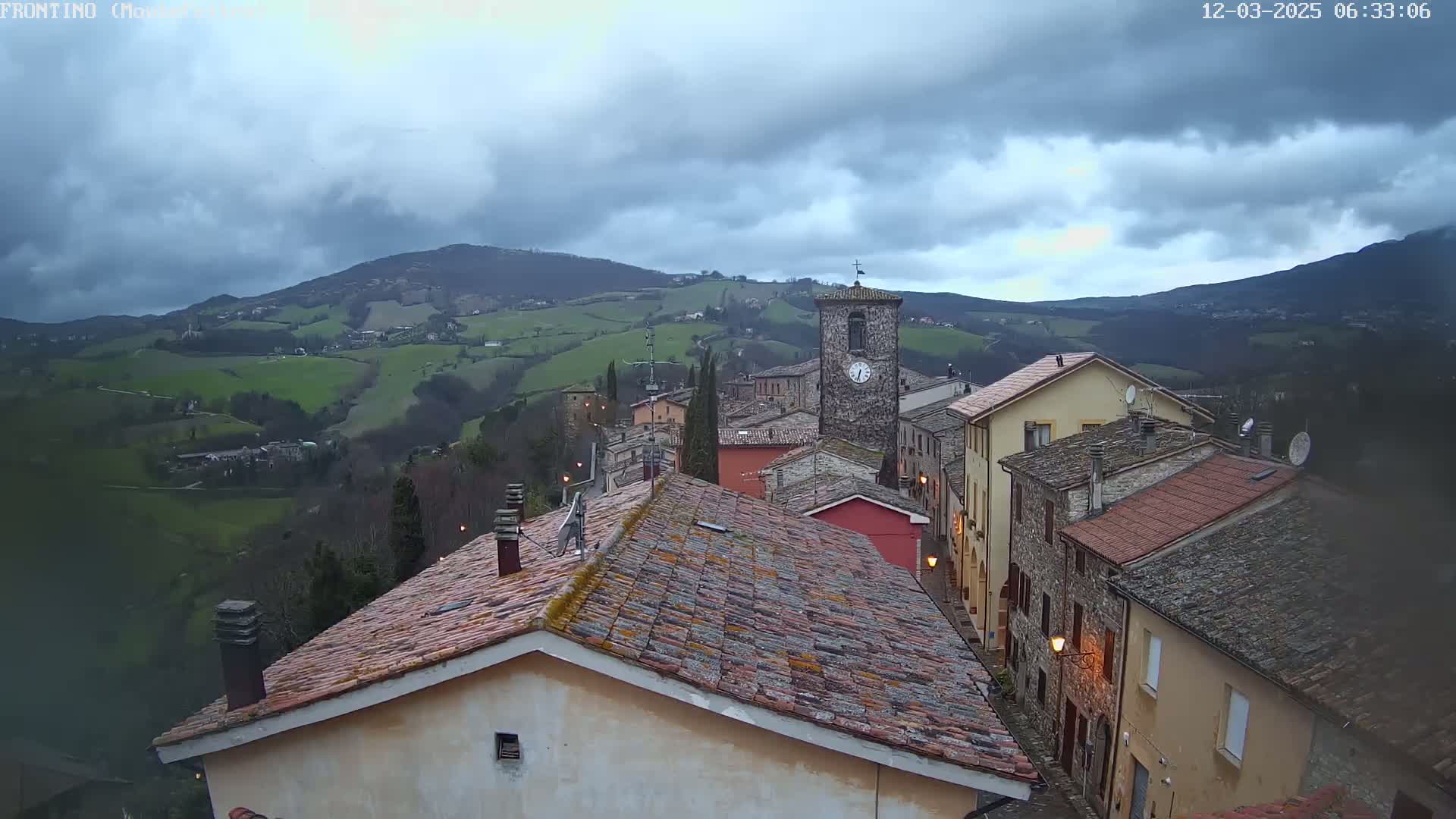 A stone village nestled in a valley under a cloudy sky, overlooks rolling green hills and distant mountains.