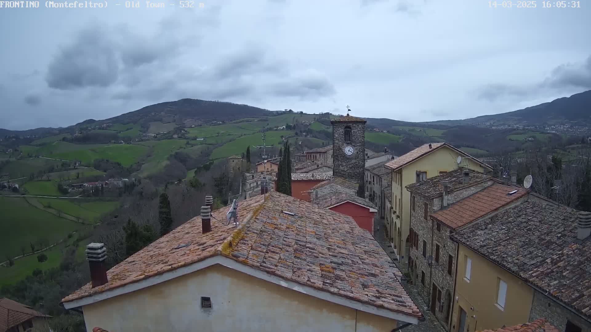 A view of a stone village nestled in rolling green hills under a cloudy sky.