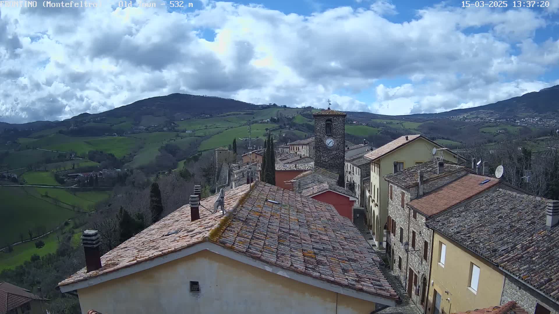 A partly cloudy day reveals a village nestled in a valley, with rolling green hills and mountains in the background.