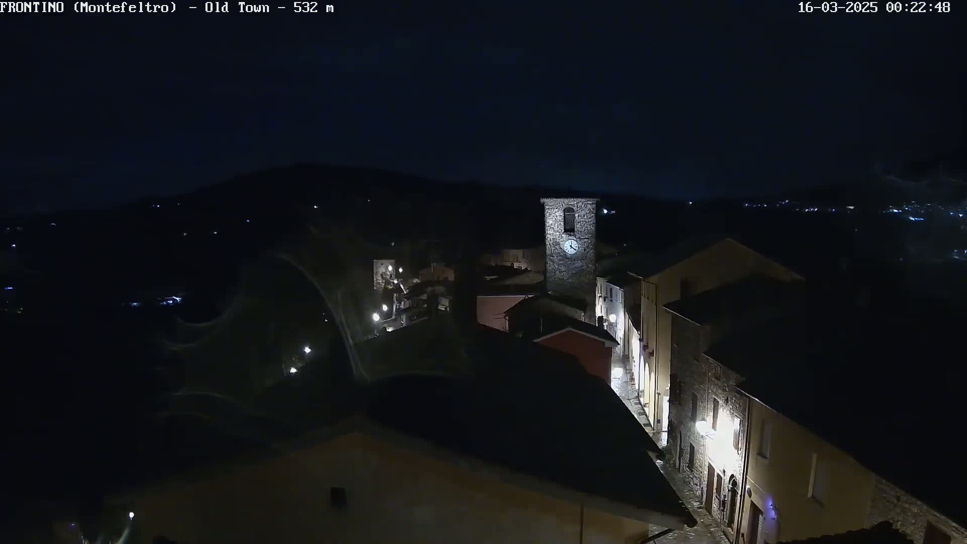 A nighttime, high-angle view of a small town with a clock tower, dimly lit streets, and buildings under a dark sky.