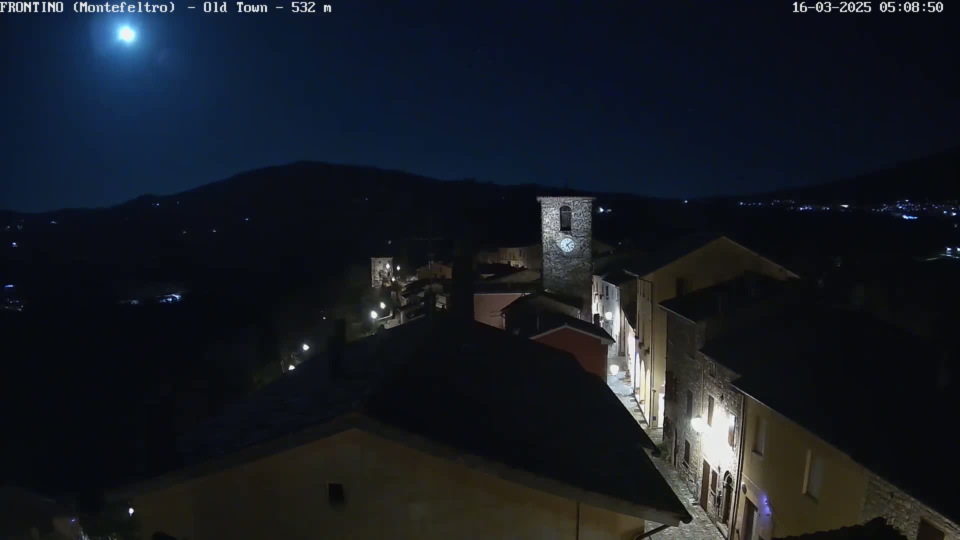 A nighttime, high-angle view of a small town nestled in a valley under a bright moon and clear sky, with streetlights illuminating the buildings and streets.
