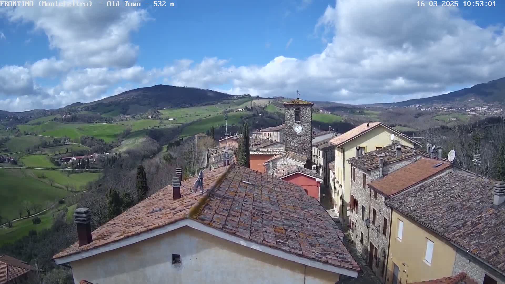A partly cloudy day reveals a quaint village nestled in rolling green hills, with terracotta-roofed buildings and a prominent clock tower.