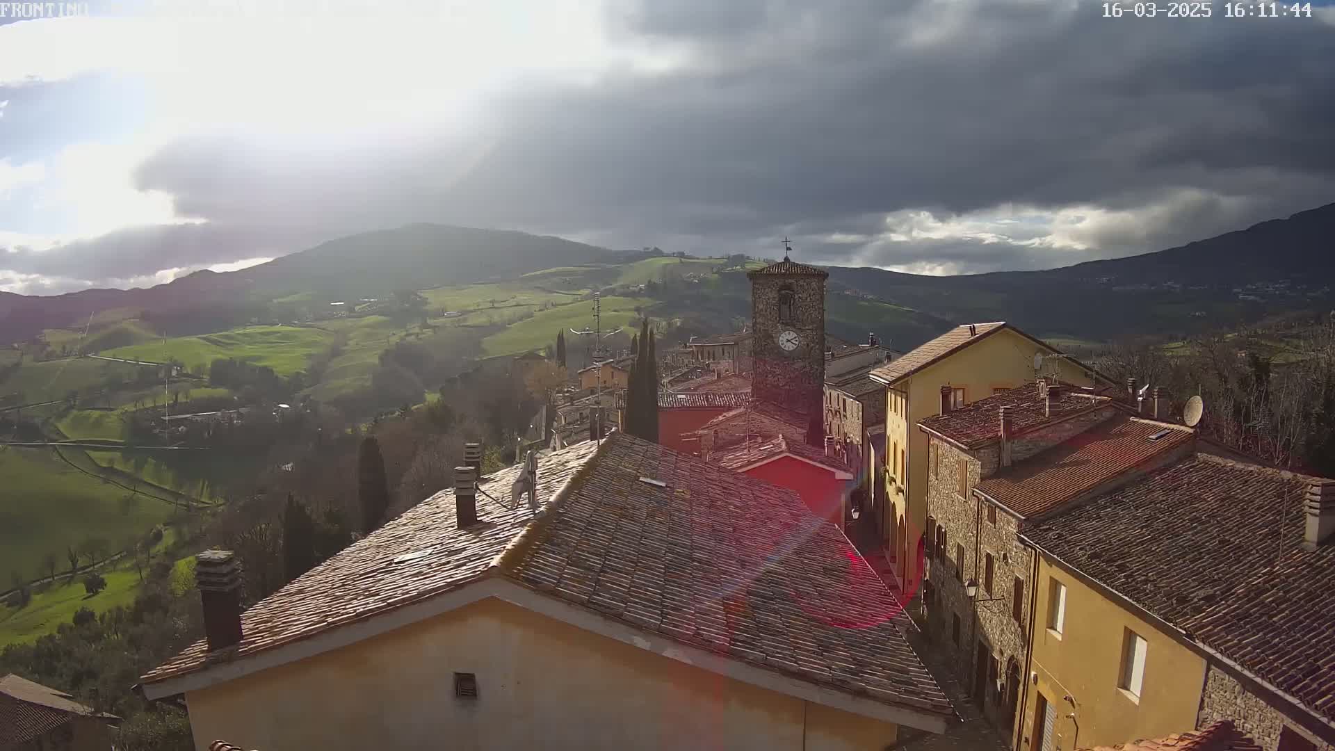 A sun-dappled view of a village nestled in rolling green hills under a partly cloudy sky.