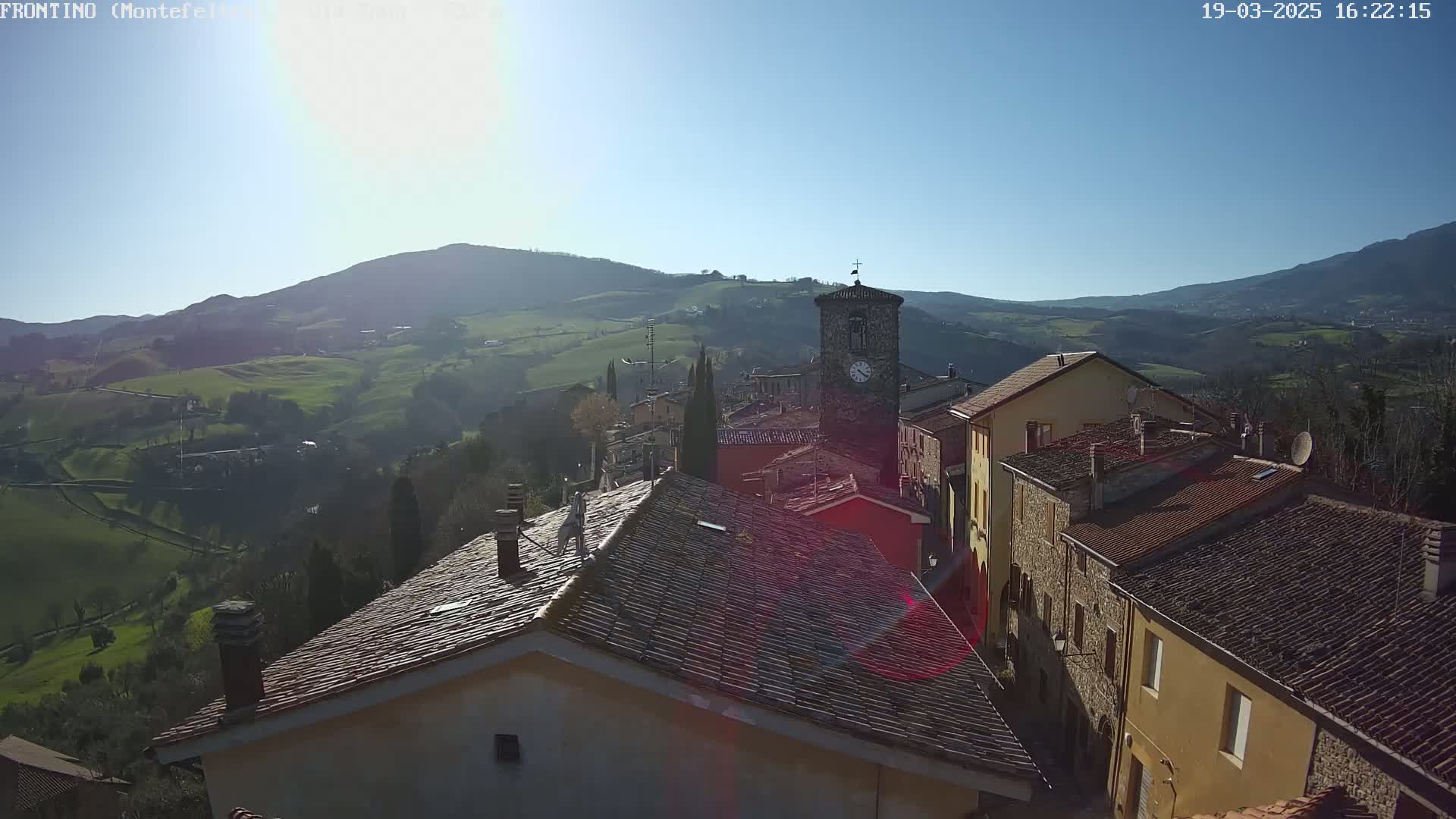 A sunlit village nestled in rolling green hills, with a clock tower visible among the stone buildings.