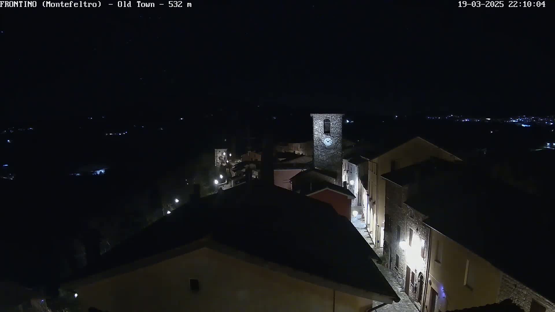 A nighttime, aerial view of a small, stone village with a clock tower, illuminated by streetlights under a clear, dark sky.
