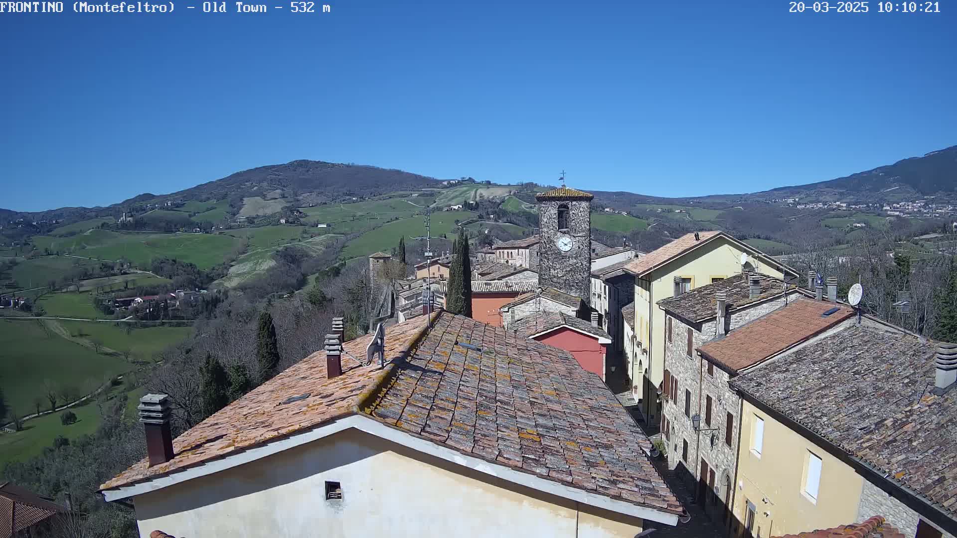 A small town nestled in a valley under a clear blue sky, with terracotta rooftops and a clock tower visible among the buildings.