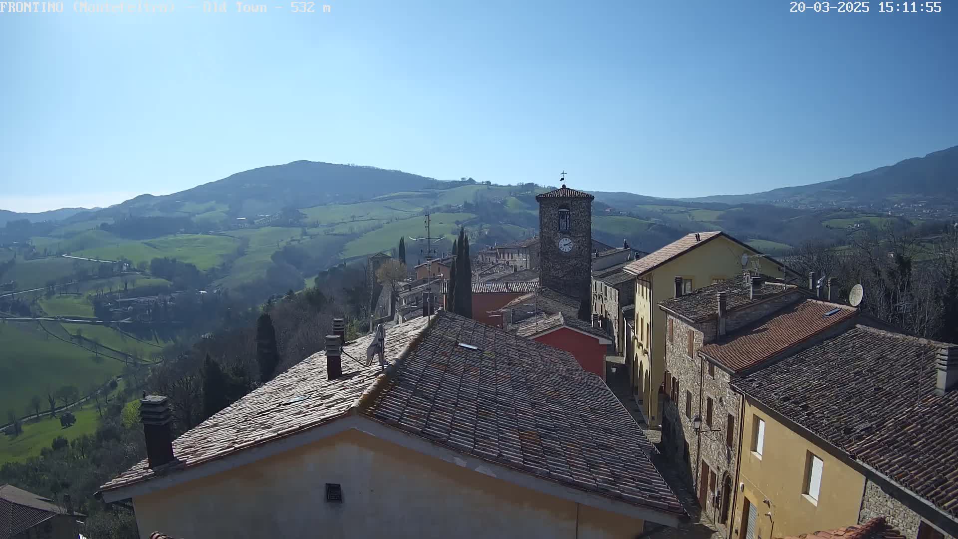 A small town nestled in a valley, under a clear blue sky, is surrounded by rolling green hills and mountains.