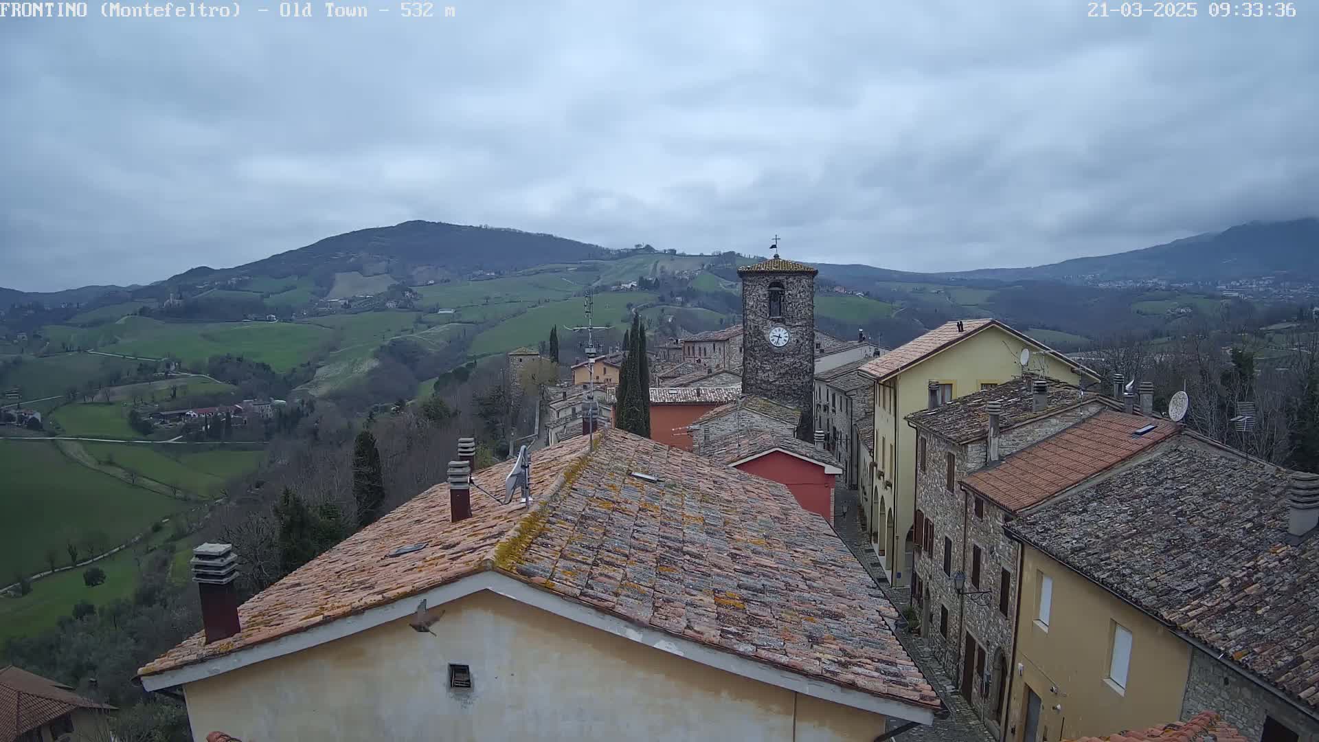 A clock tower is visible in the center of a stone village nestled in a valley, under an overcast sky, with rolling green hills in the background.