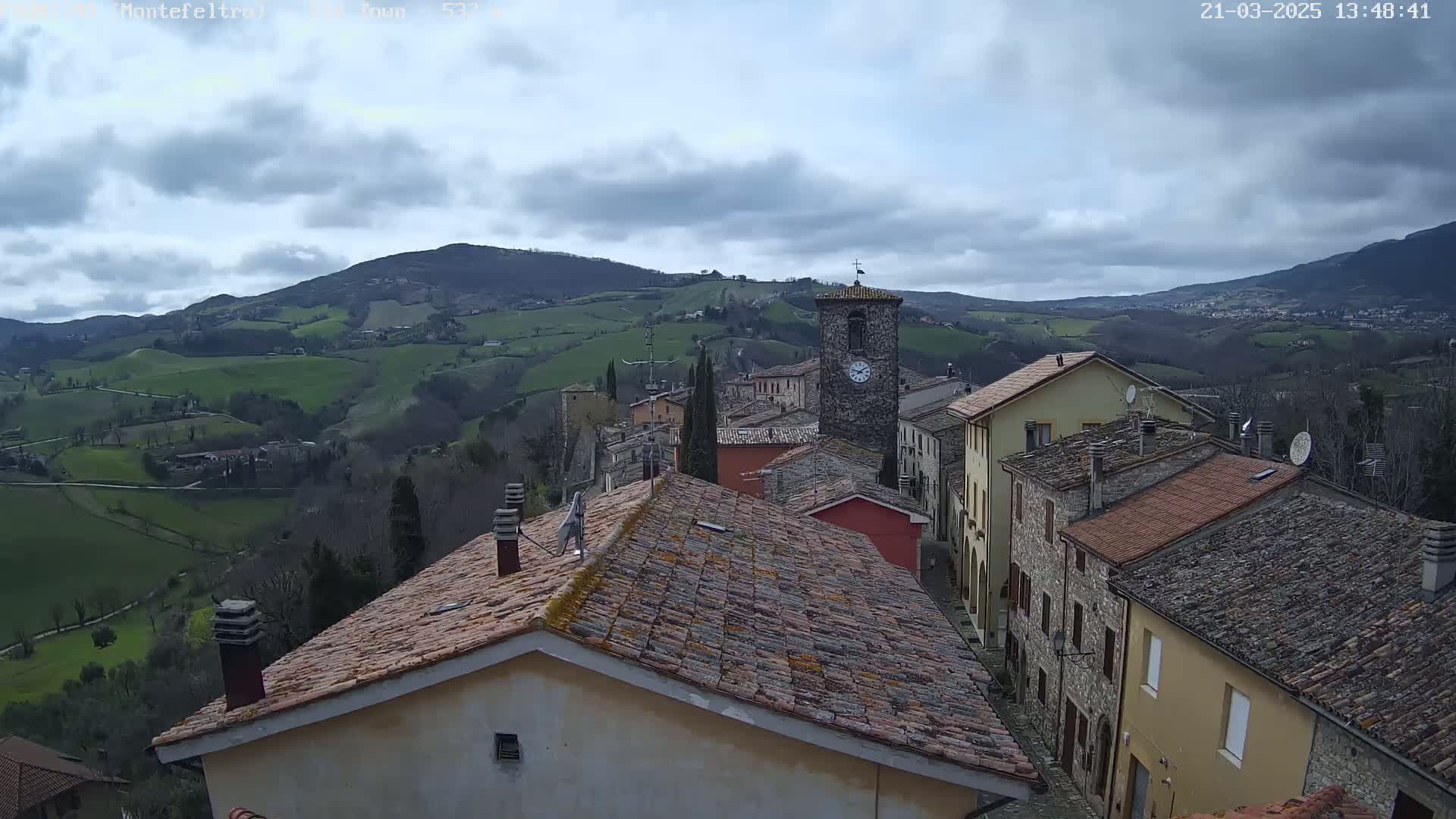 A stone village nestled in a valley, under a cloudy sky, is surrounded by rolling green hills.