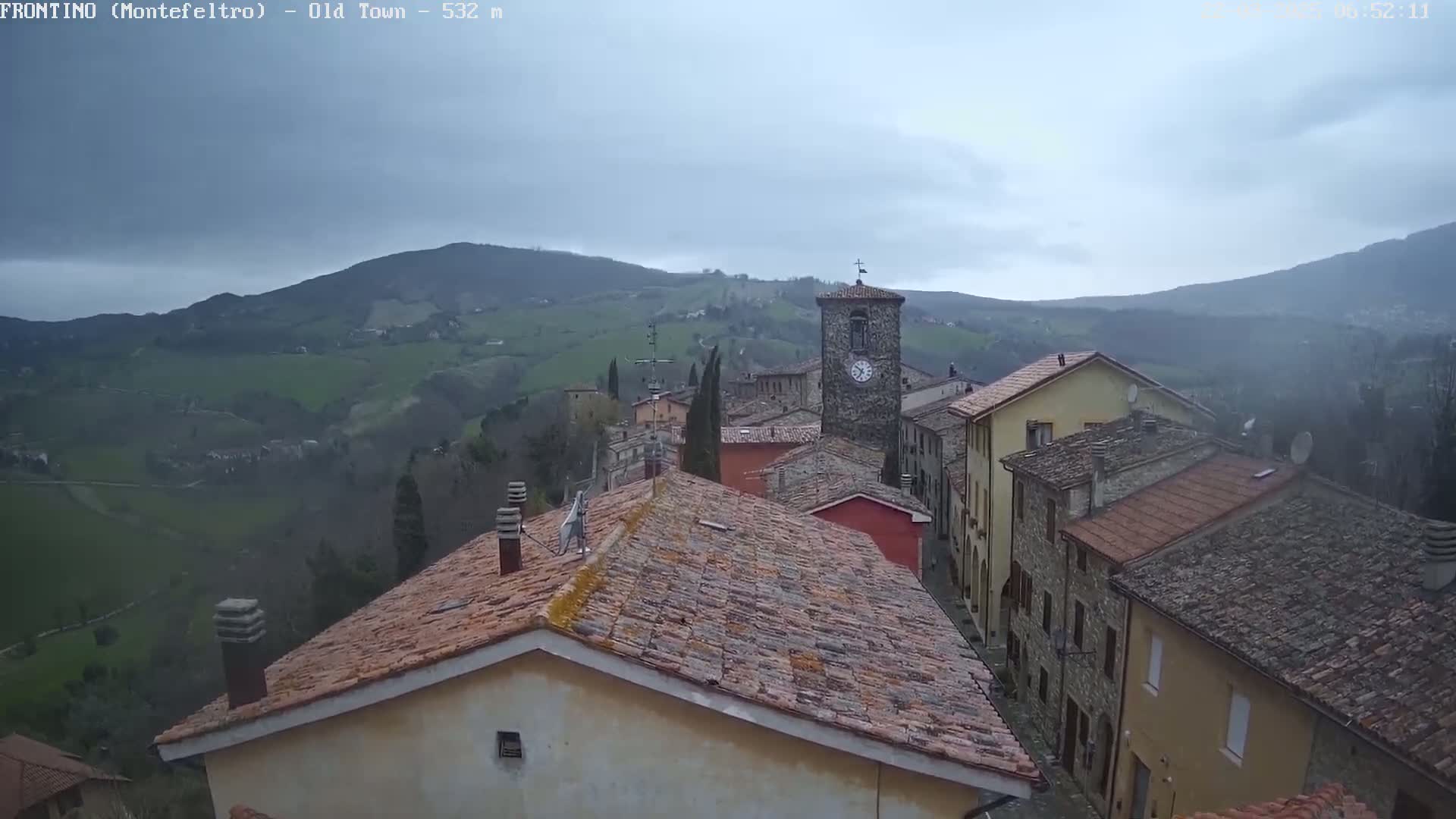A stone village with a clock tower is nestled in a valley, overlooked by rolling green hills under a cloudy sky.