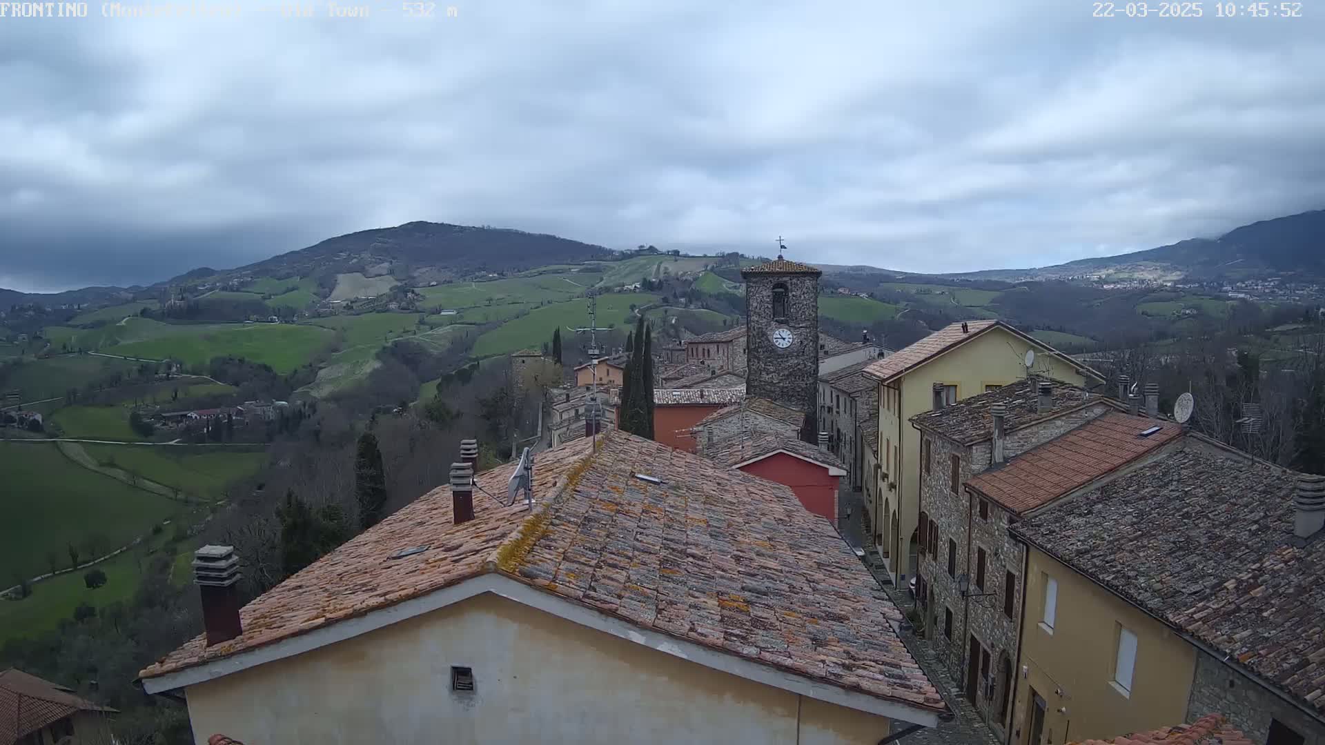 A quaint village nestled in rolling green hills under a cloudy sky, with a clock tower prominent among the stone buildings.