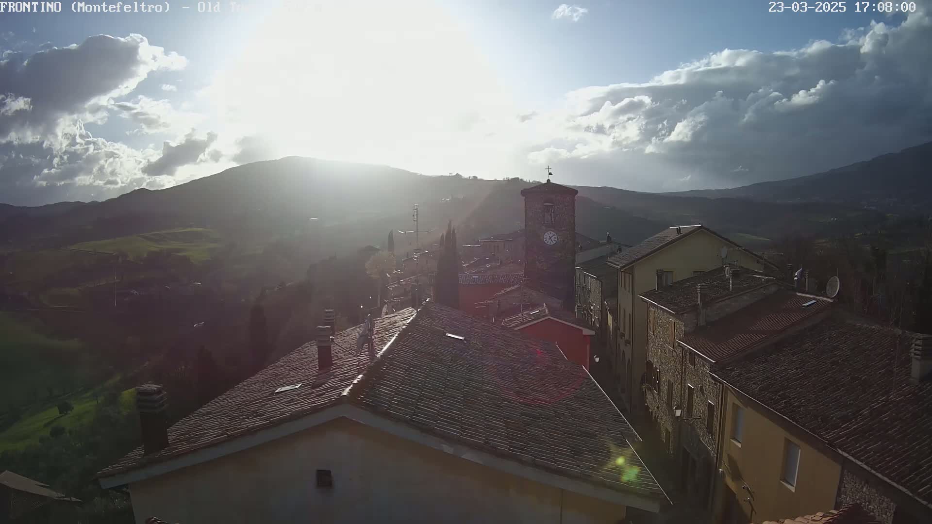 A sunlit village nestled in a valley between hills, under a partly cloudy sky.
