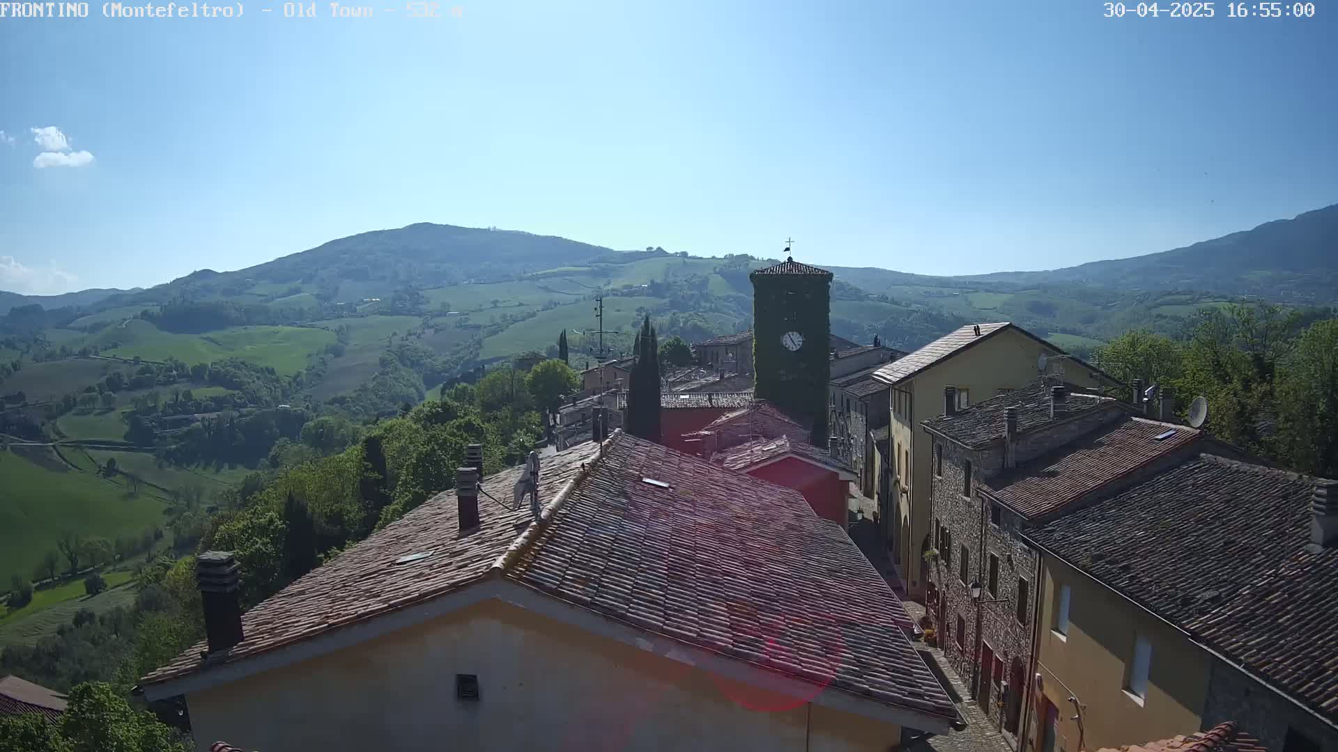 A quaint village nestled in a valley, with rolling green hills and a clear blue sky, is seen from a high vantage point.