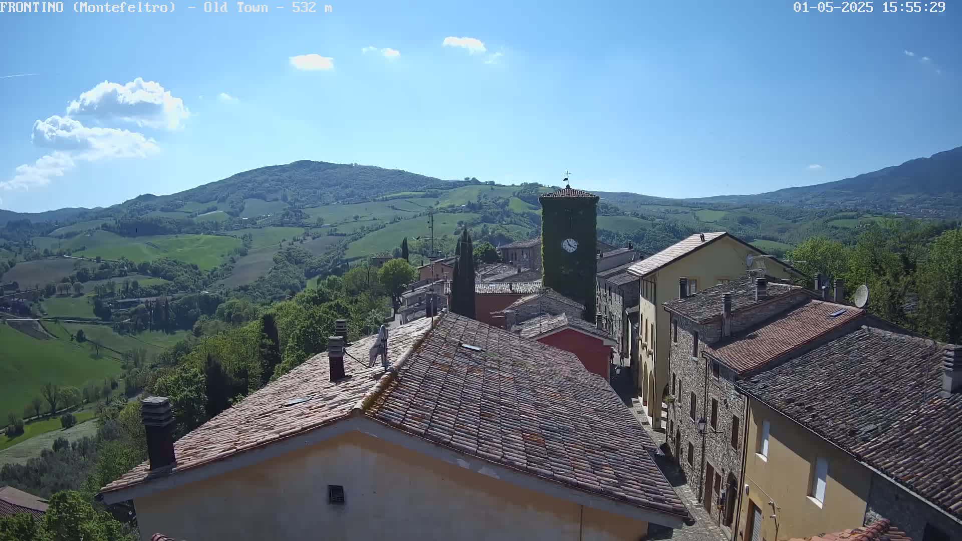 A quaint village nestled in a valley under a mostly sunny sky, with rolling green hills visible in the background.