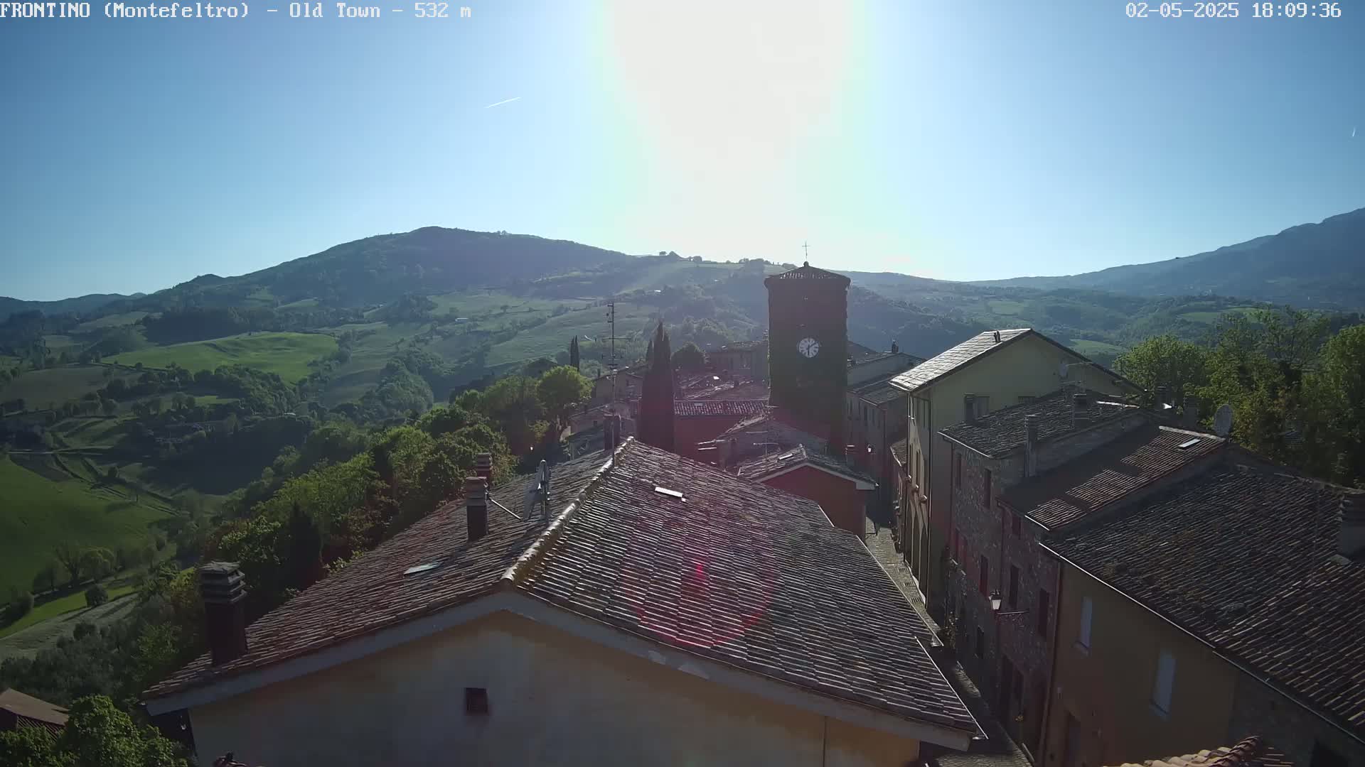 A sunlit view of a small town nestled in a verdant valley, with rolling hills and mountains in the background under a clear blue sky.