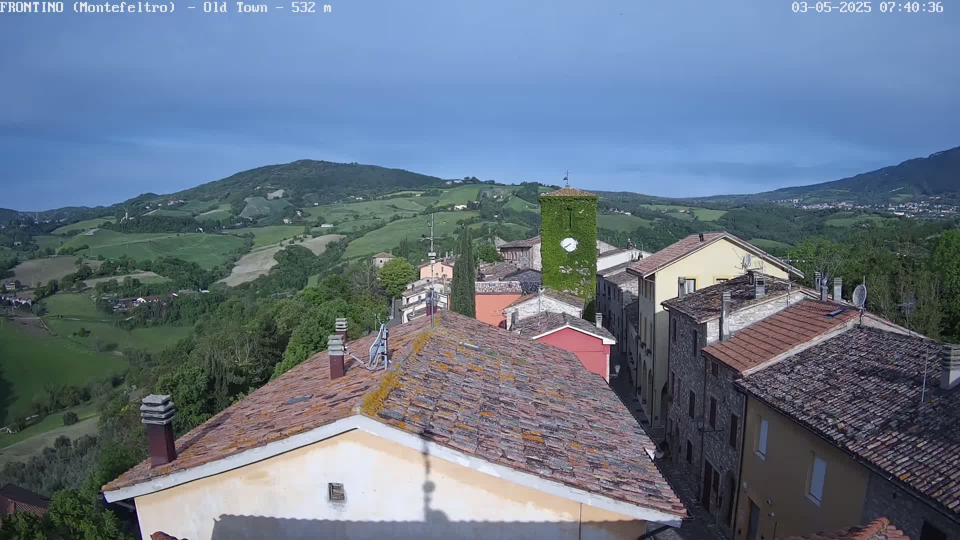 A view of a small town nestled in a valley under a mostly clear blue sky, with rolling green hills in the background.