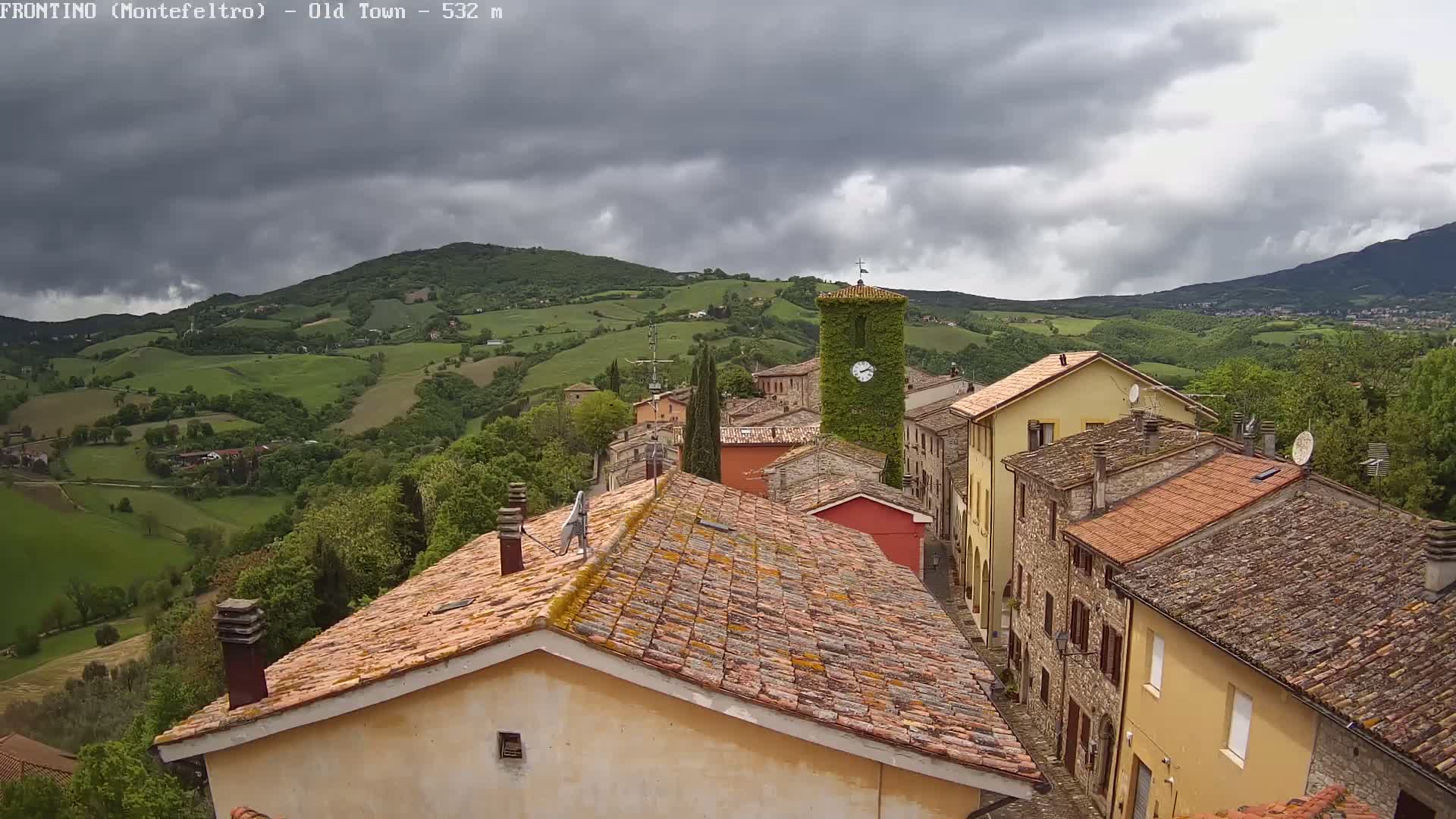 A small town with terracotta roofs nestles in a green valley under a cloudy sky.