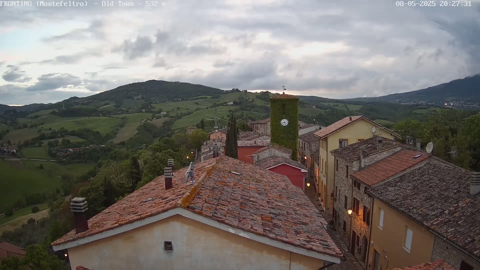 A hilltop village with terracotta rooftops and a clock tower is nestled in a valley under a cloudy twilight sky.