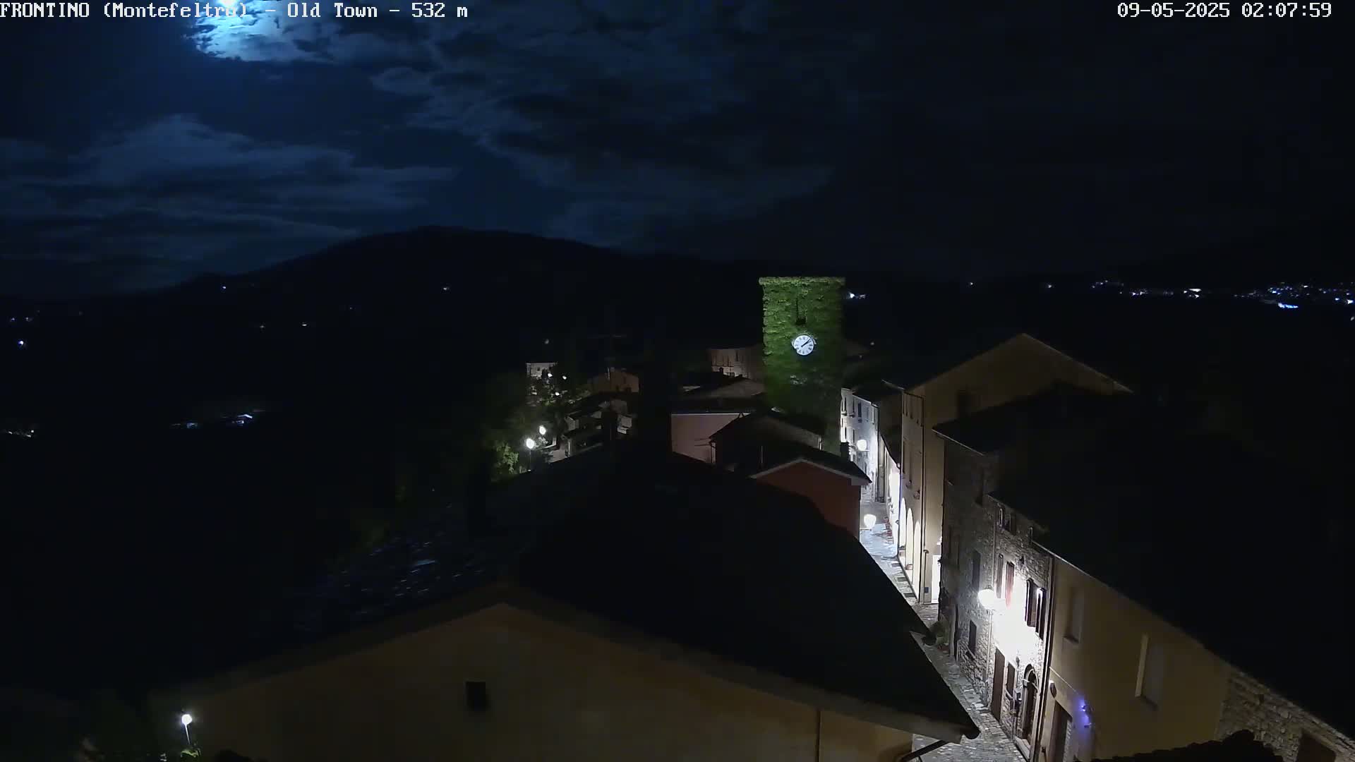 A nighttime, high-angle view of a village street with dimly lit buildings under a partly cloudy sky.